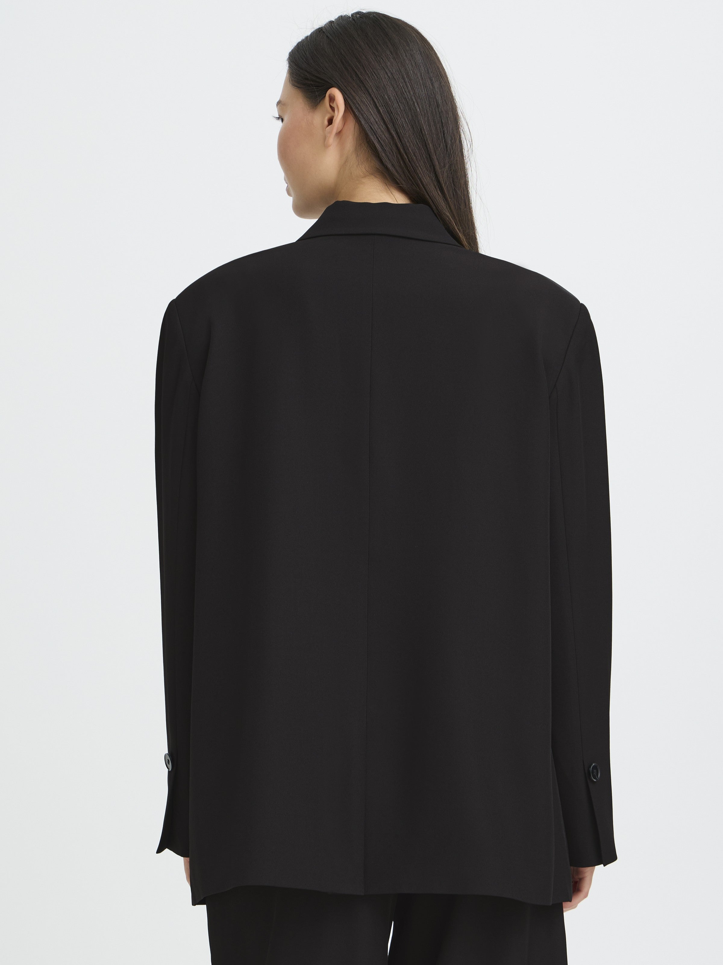 Back view of a black oversized jacket with long sleeves and buttoned cuffs on a person with long dark hair.