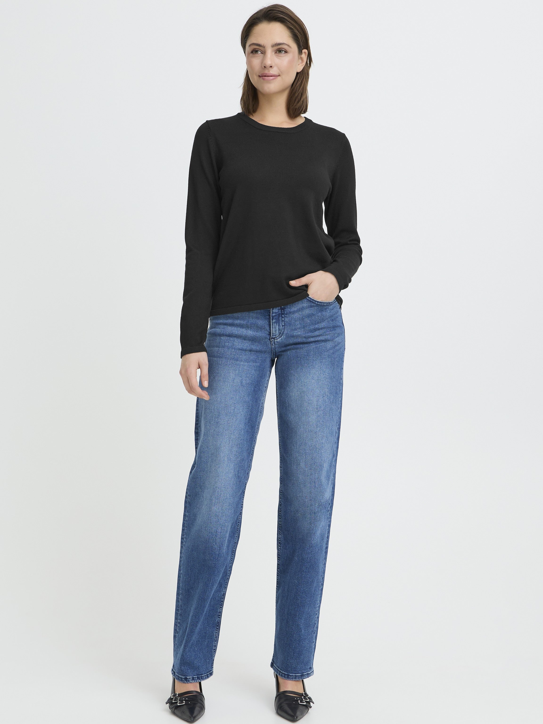 Individual standing in blue denim jeans, black long-sleeve top, and black pointed shoes against a plain background.