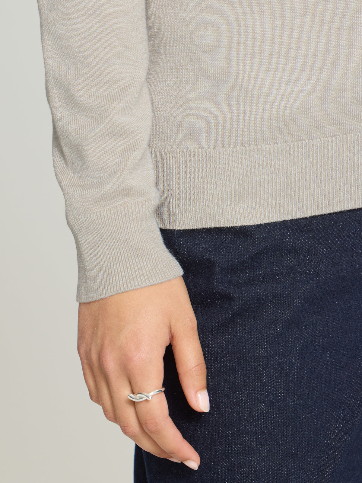 Close-up of a light beige sweater sleeve and hand with a silver ring over dark pants fabric.