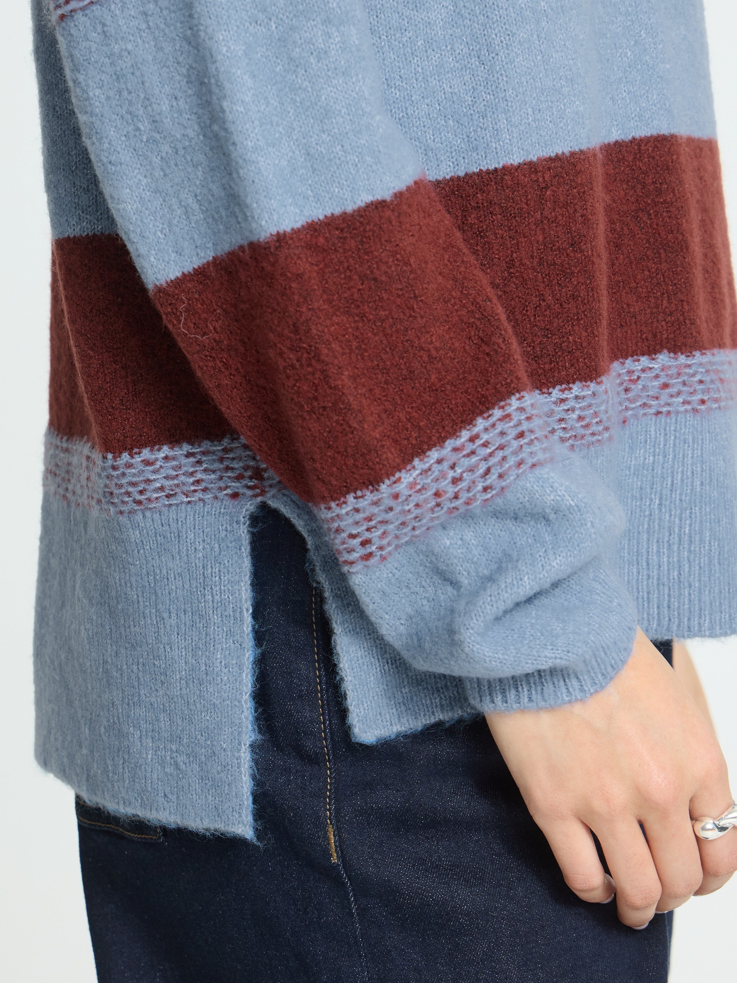 Close-up of a blue and rust striped wool sweater sleeve and side slit detail paired with dark denim pants.