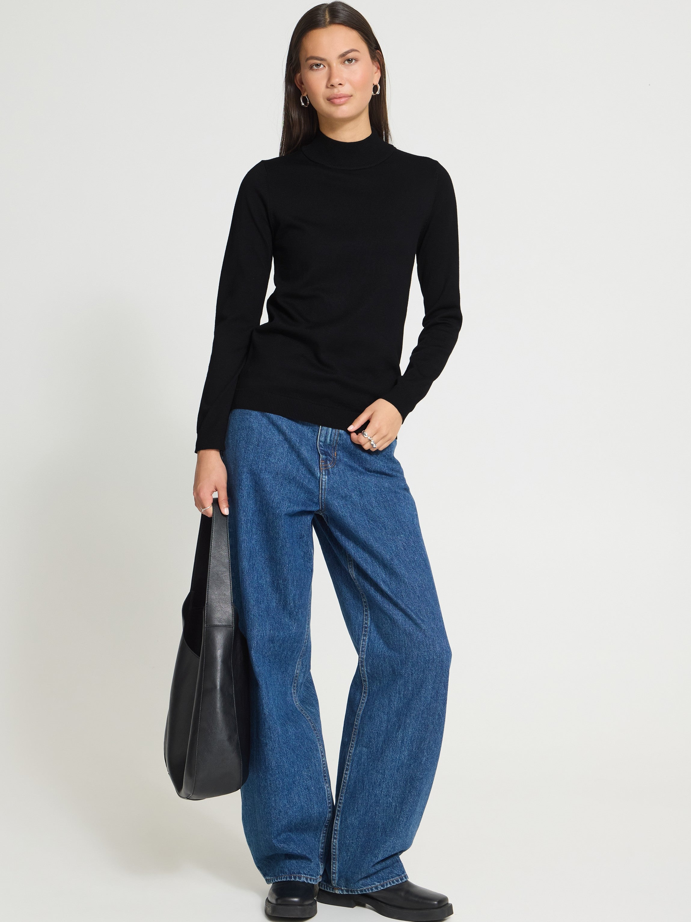 Individual in a black long-sleeve top and blue wide-leg denim jeans holding a large black leather bag