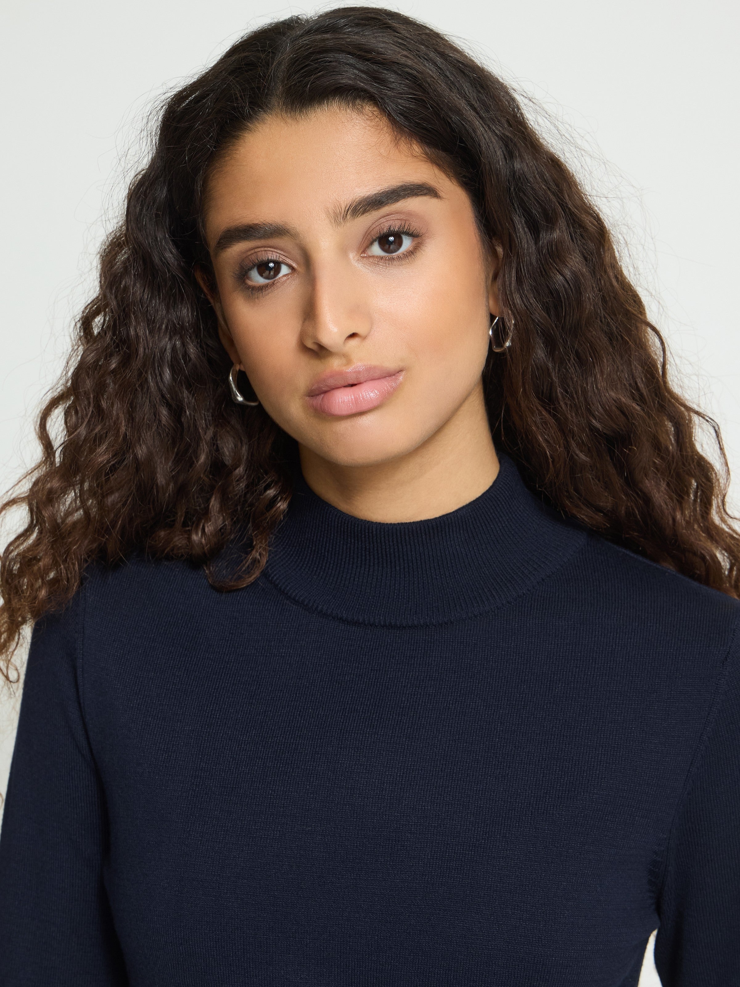 Close-up of curly dark hair and hoop earrings with a navy blue high-neck sweater on a grey background.