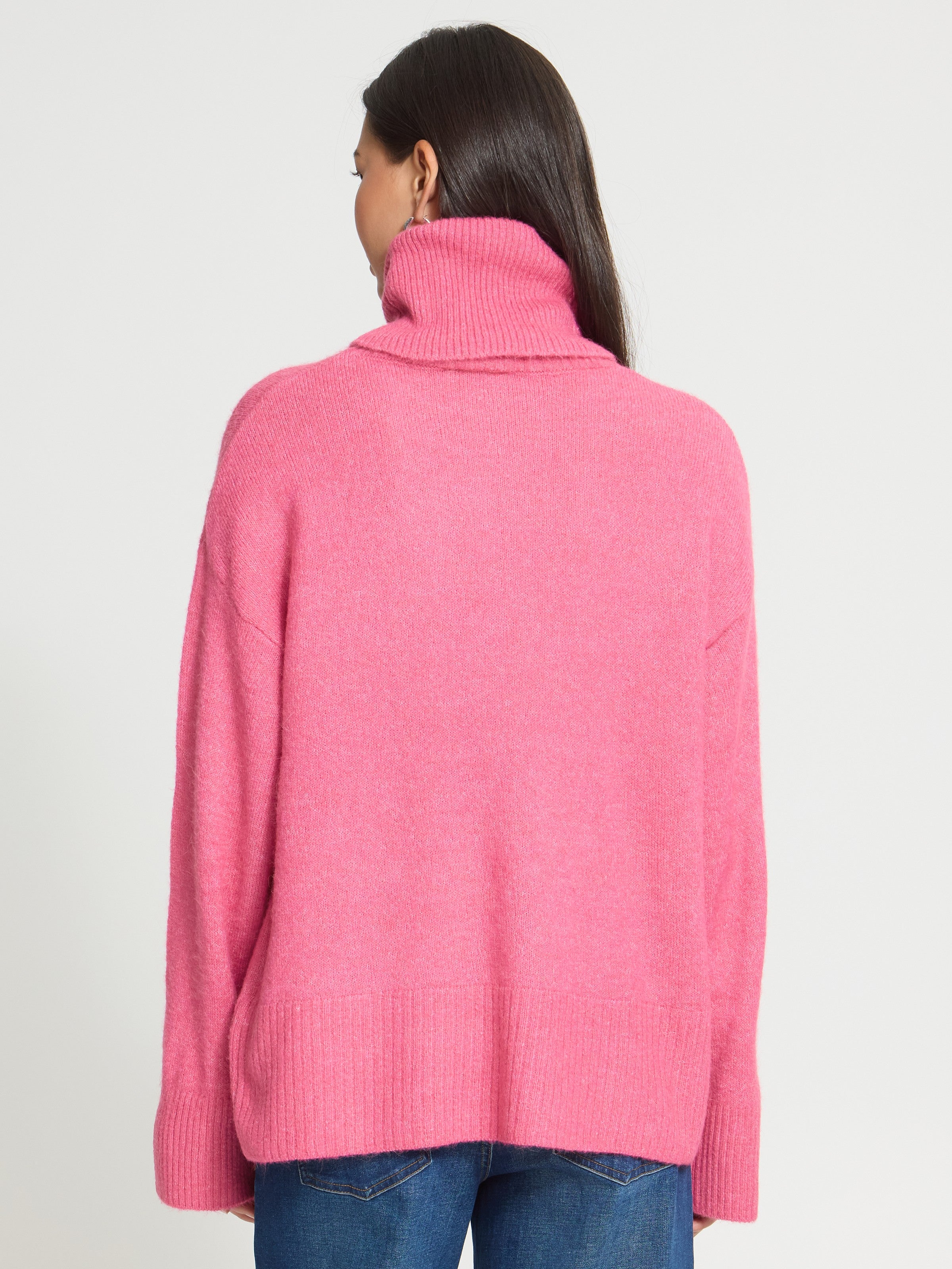 Back view of a bright pink turtleneck sweater with ribbed cuffs and hem paired with blue denim jeans.