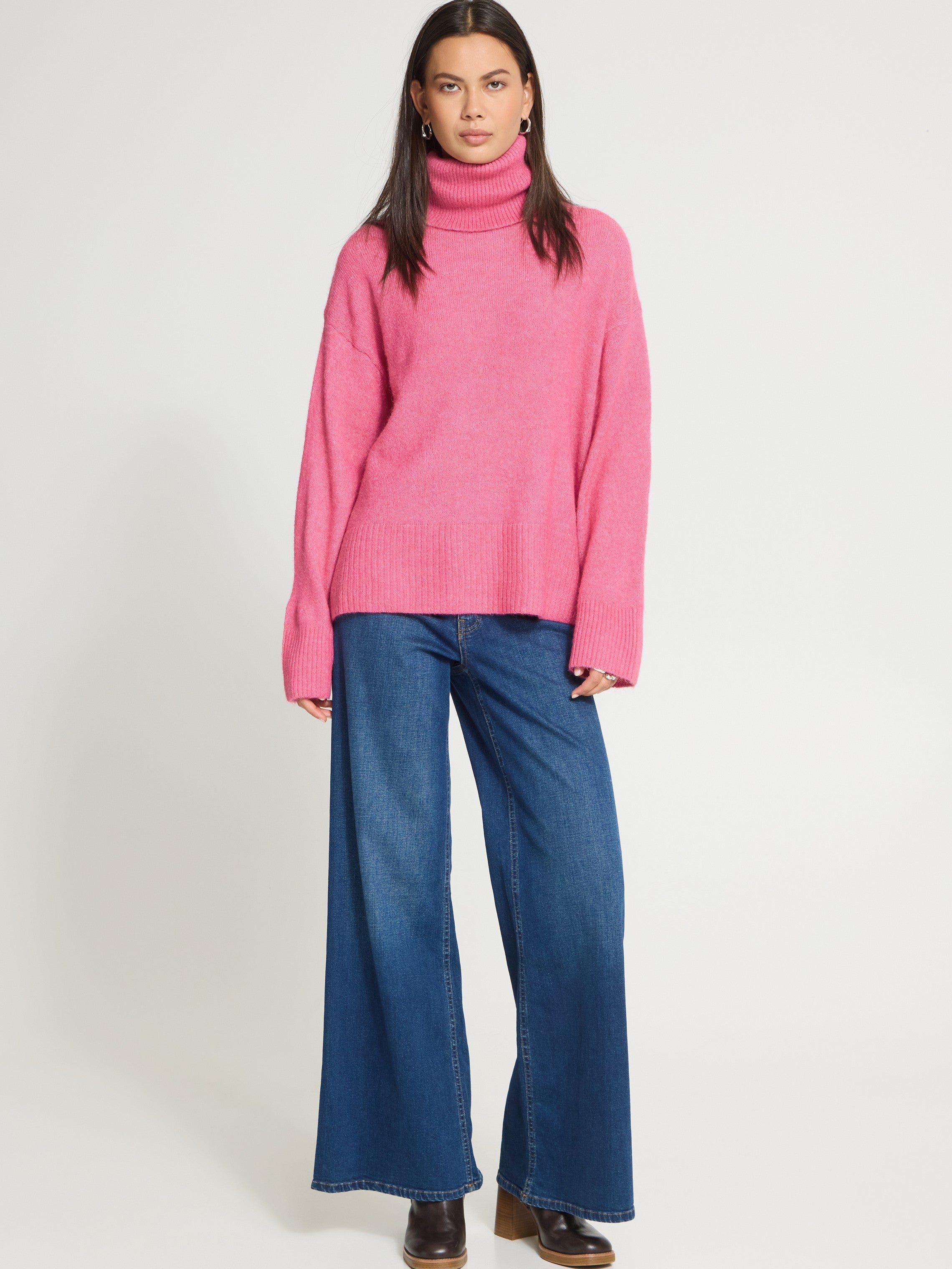 Model in a bright pink turtleneck sweater paired with wide-leg blue denim jeans and brown shoes against a plain background.