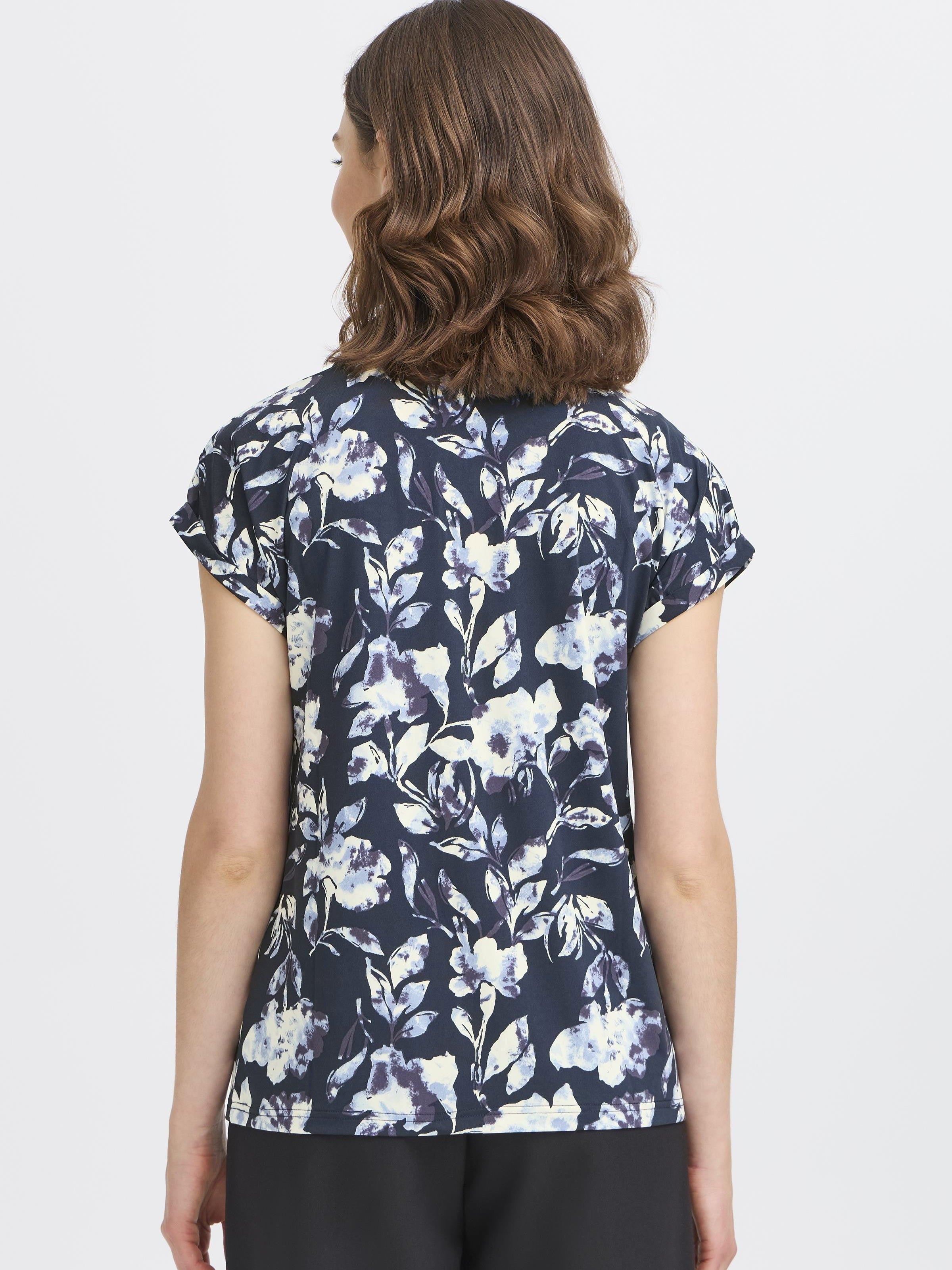 Back view of a dark floral Fransa Seen Short Sleeve Crew Neck Top on model with wavy brown hair and black pants