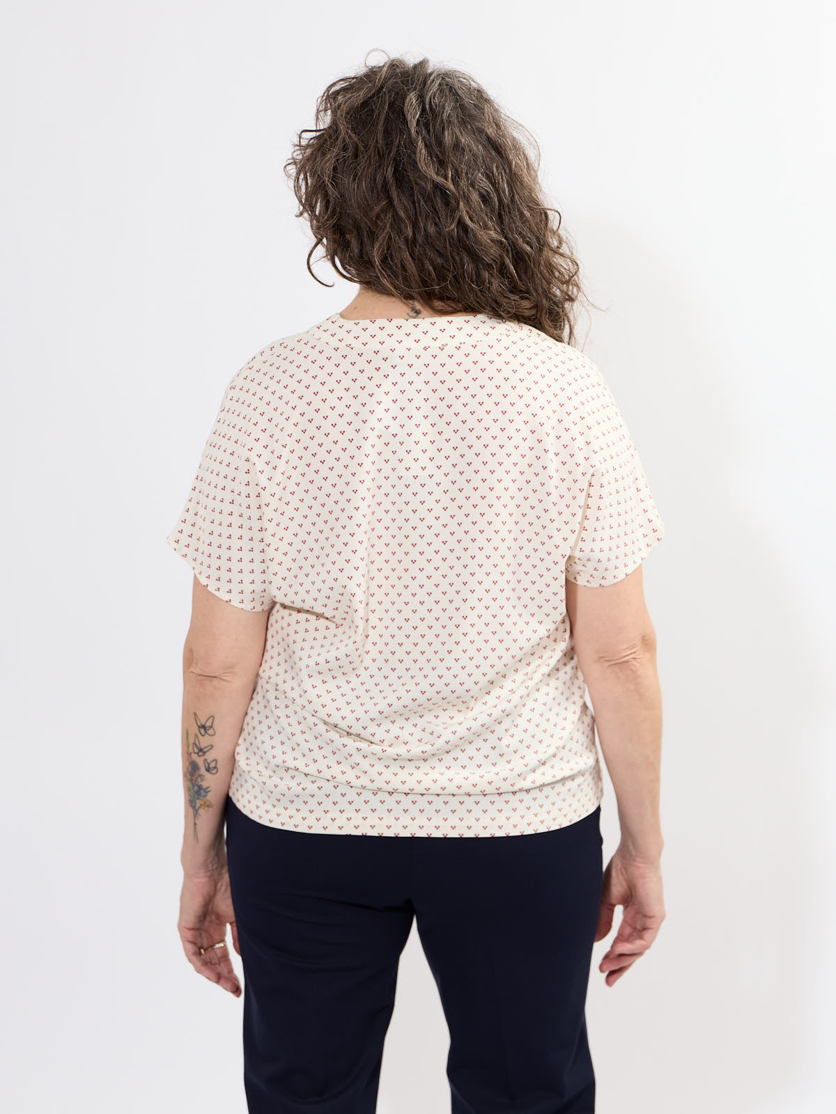 Back view of Fransa Justine Heart Print Short Sleeve V-Neck Top in off-white with small red heart prints, paired with dark pants.
