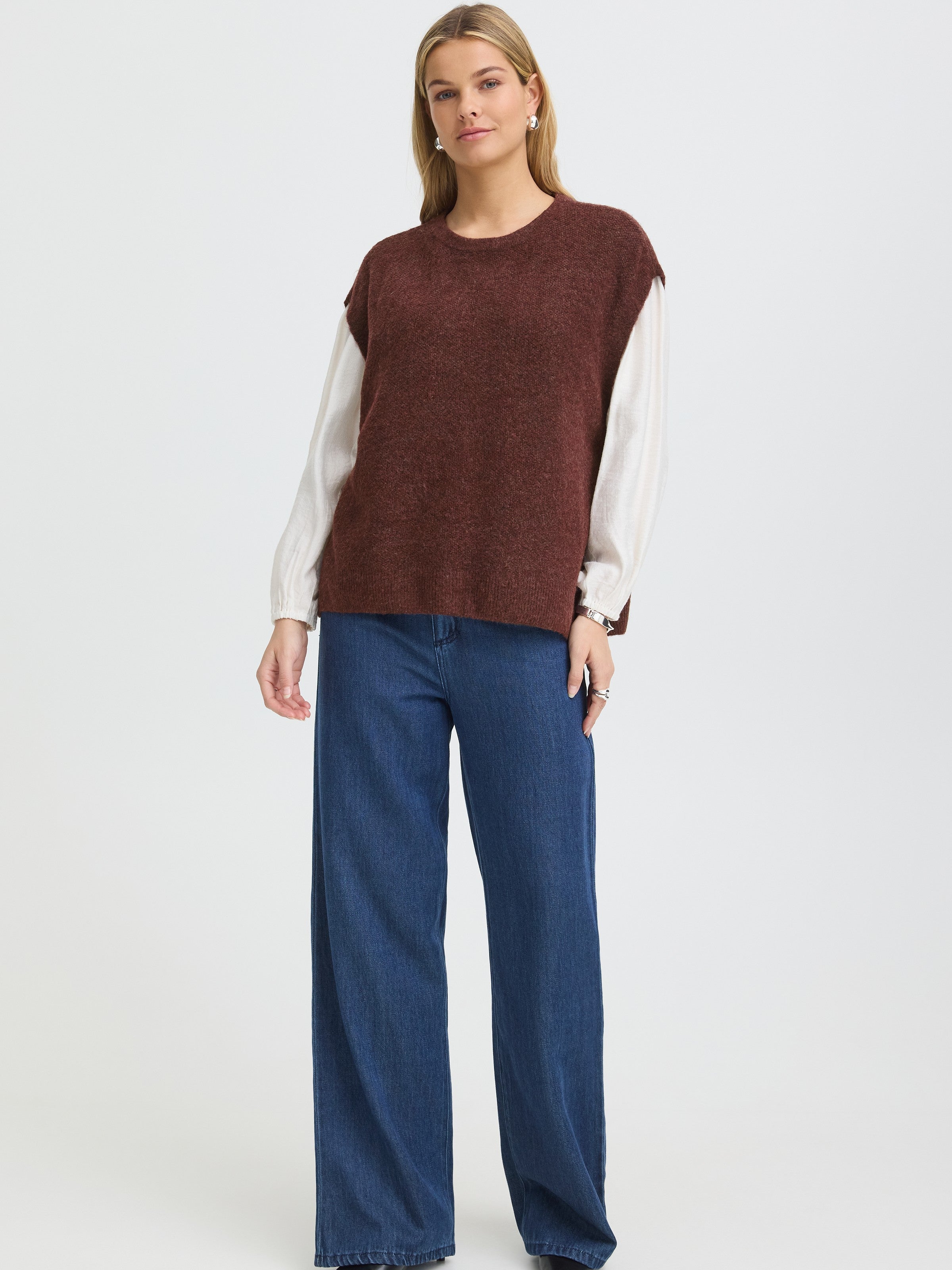 Model wearing brown sleeveless sweater over white long-sleeve top paired with wide-leg blue denim pants