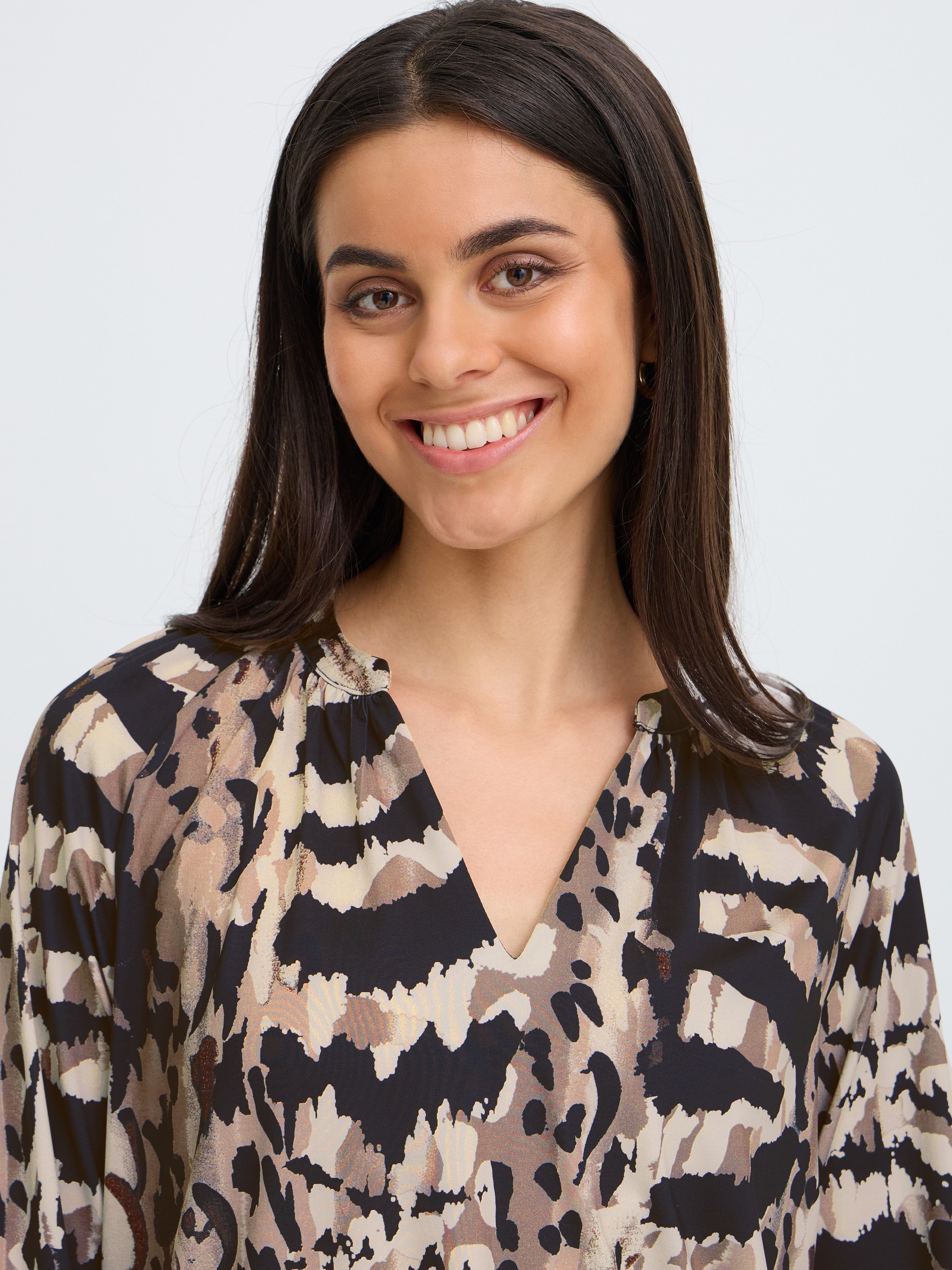 Close-up of a smiling individual with long dark hair and a patterned beige, black, and brown top with a small collar.