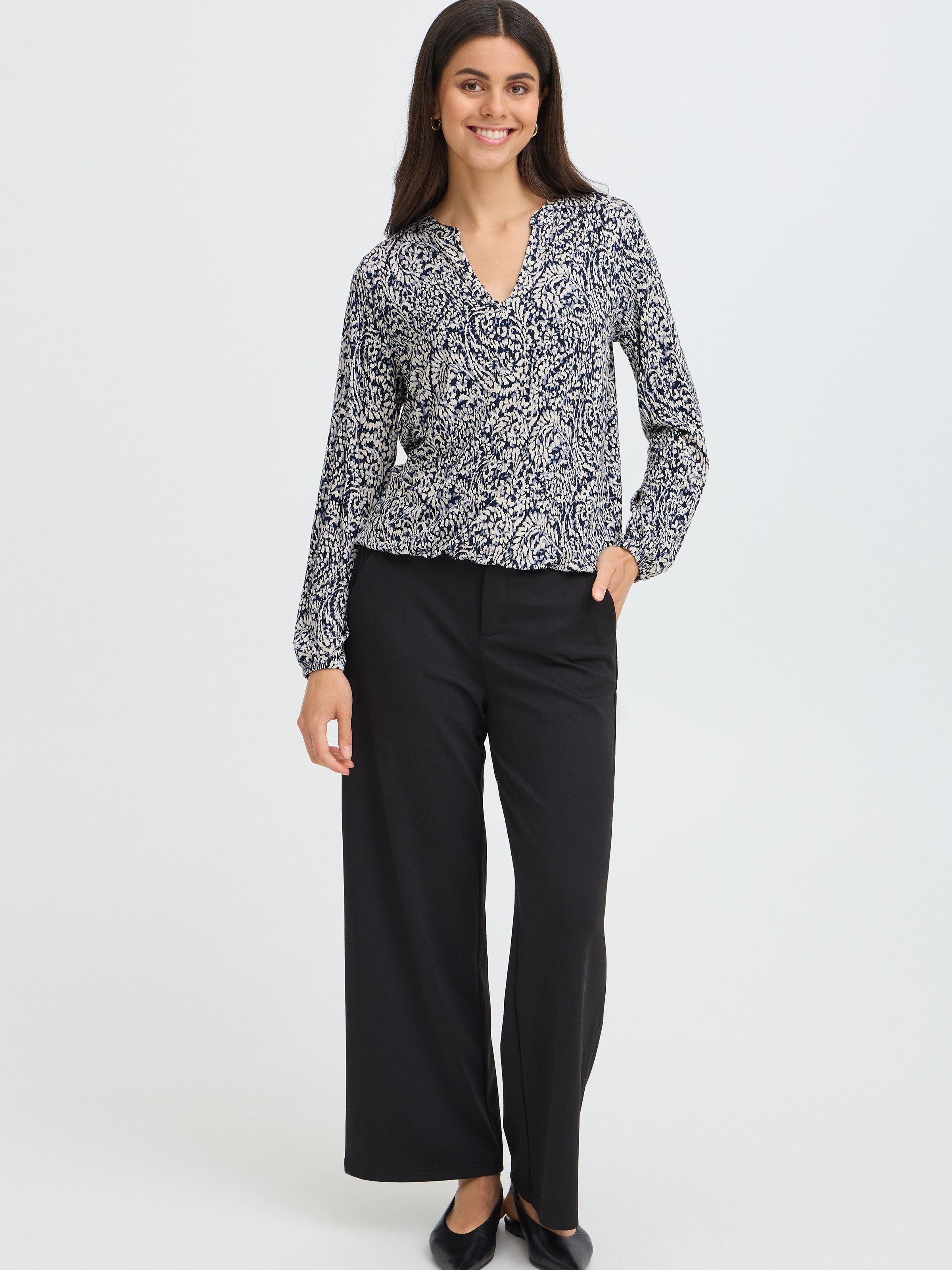 Smiling figure standing against a plain background, dressed in a patterned long-sleeve top and black wide-leg pants.