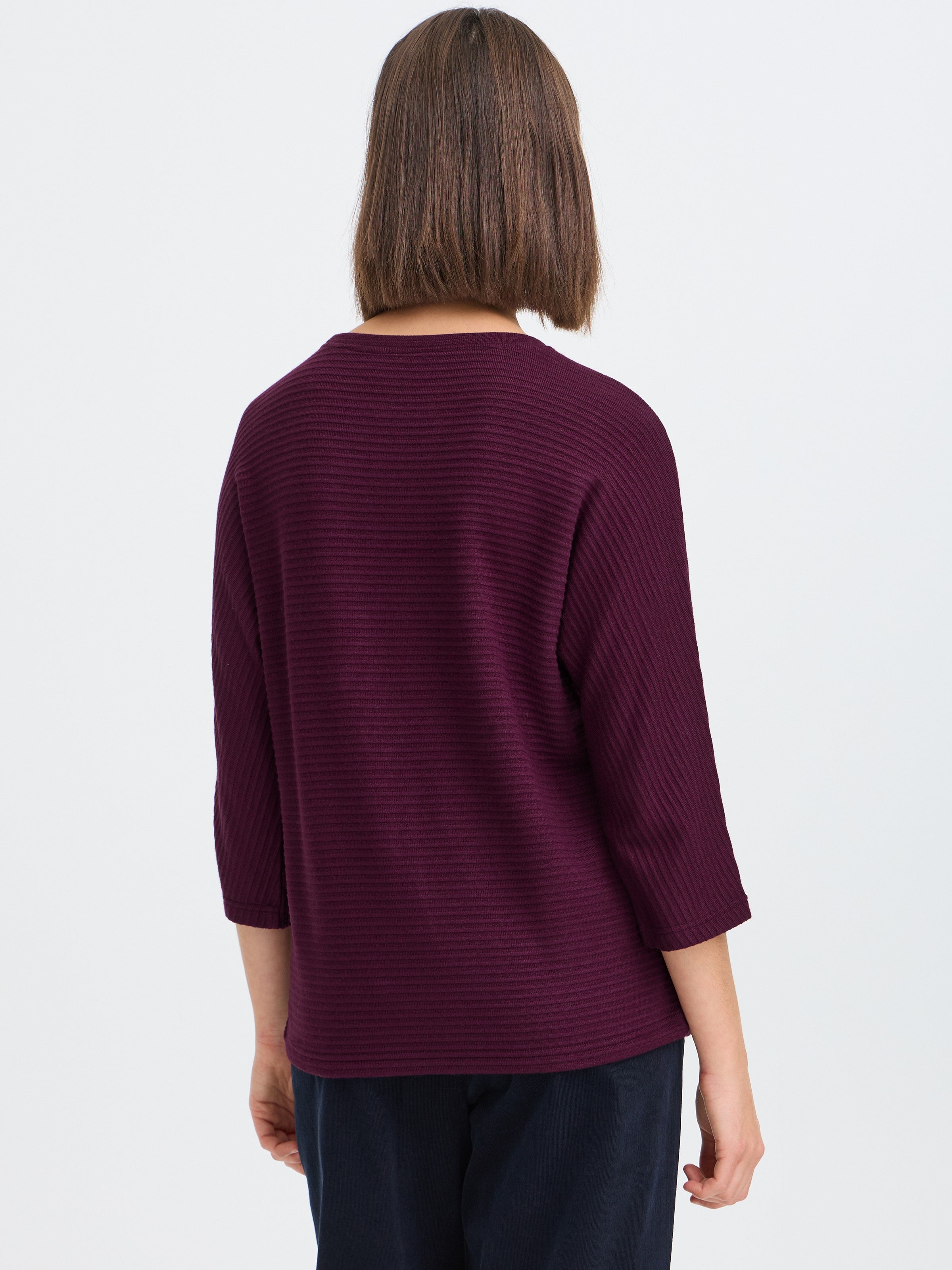 Back view of a ribbed long sleeve top in deep purple paired with dark pants against a plain light background.