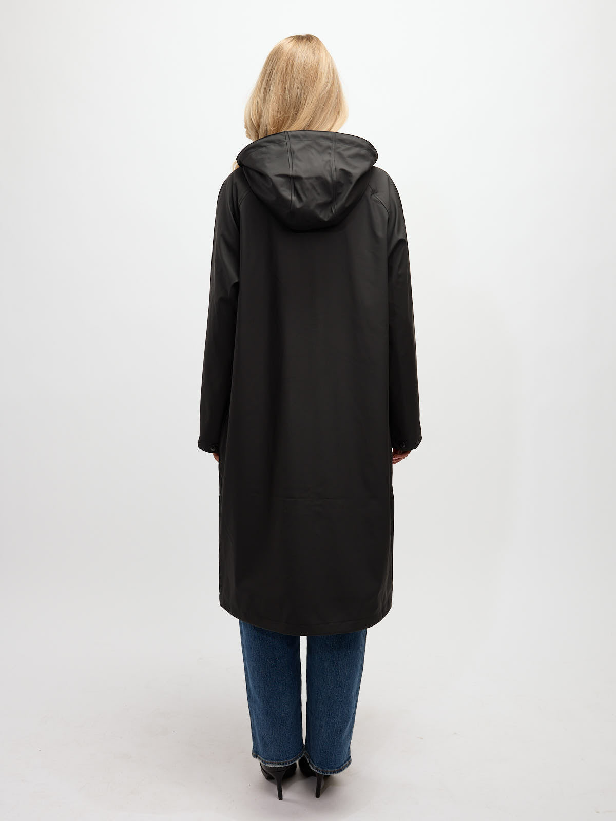 Back view of a person wearing a long black hooded coat paired with blue jeans and black heels against a plain background.