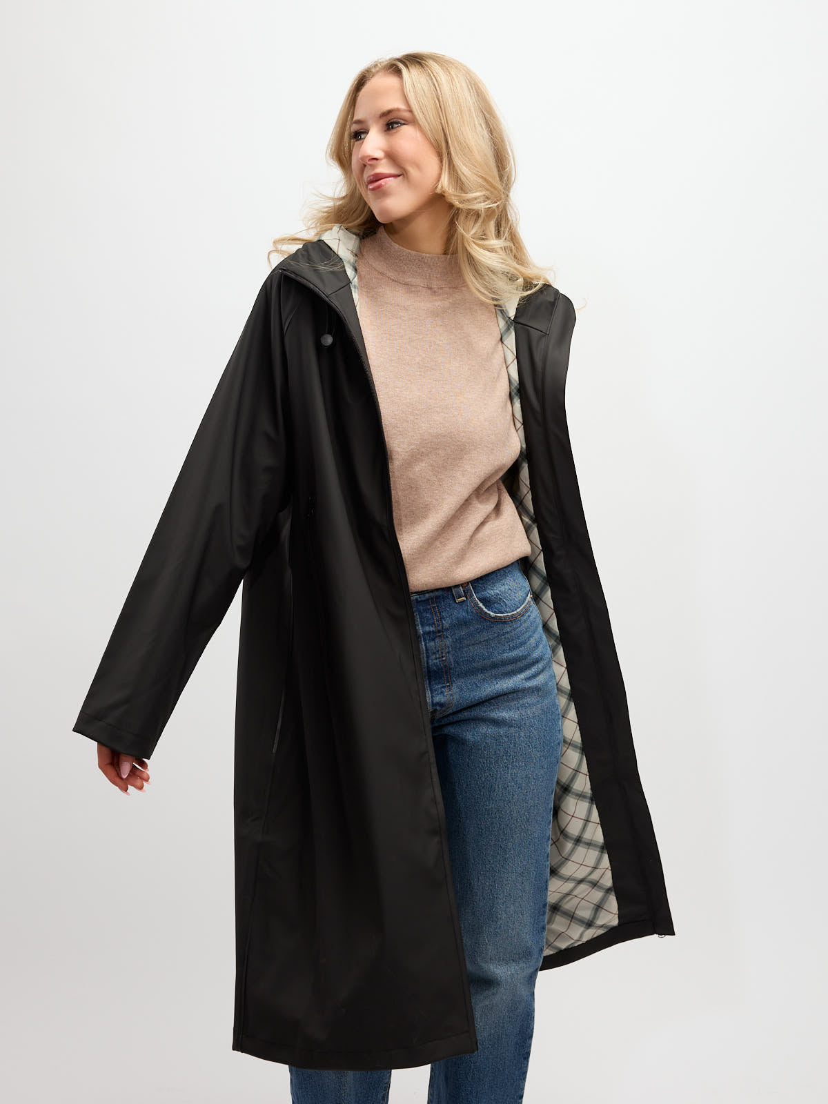 Person wearing a long black raincoat with a plaid interior lining over a beige sweater and blue jeans