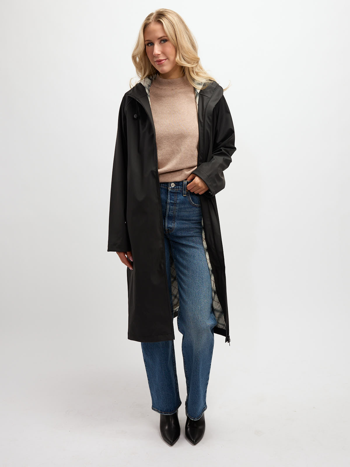 Person in a long black hooded raincoat over a beige sweater and blue jeans with black ankle boots on white background