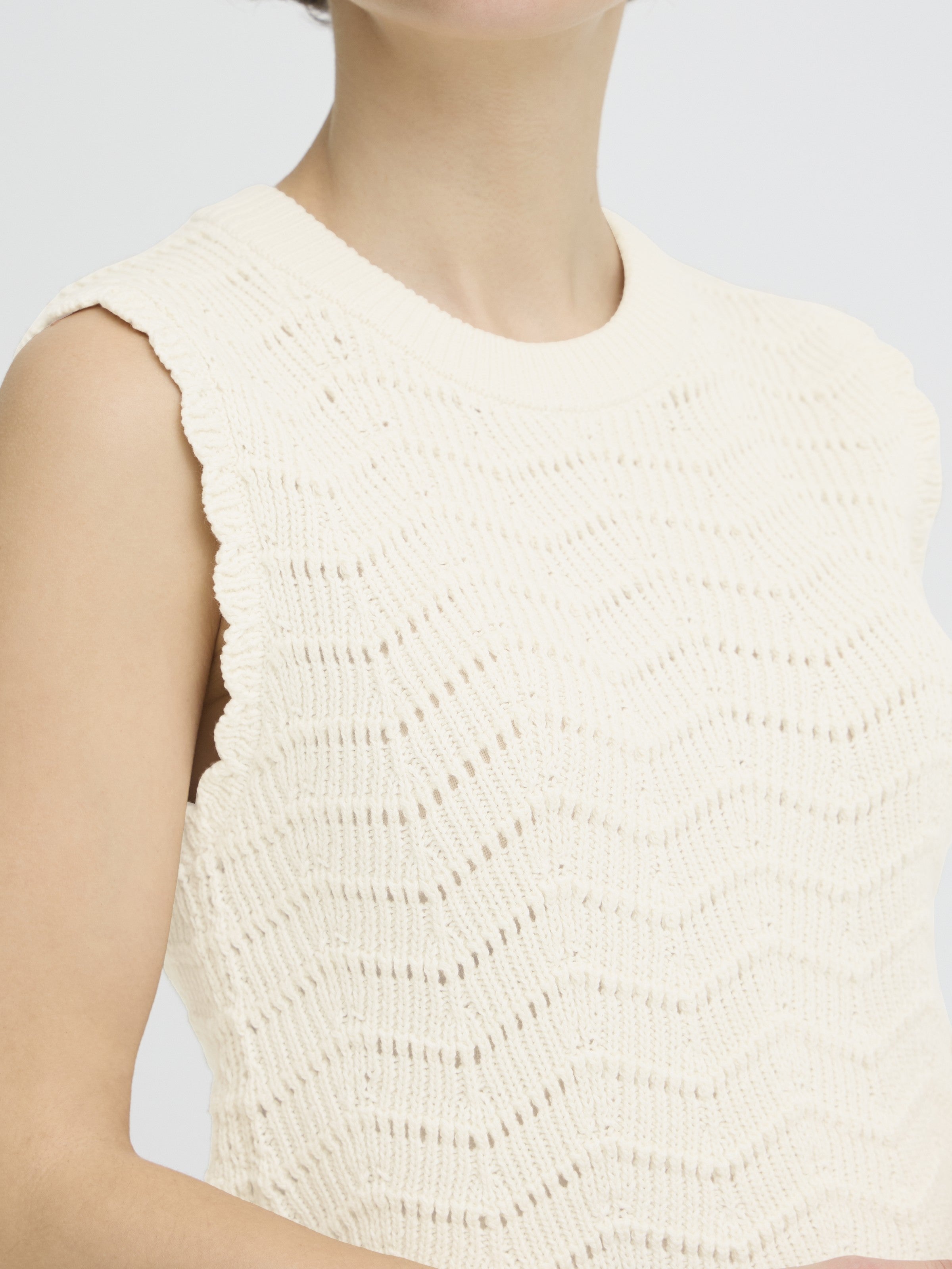 Close-up of a beige knitted sleeveless sweater with a textured zigzag pattern and scalloped edges.