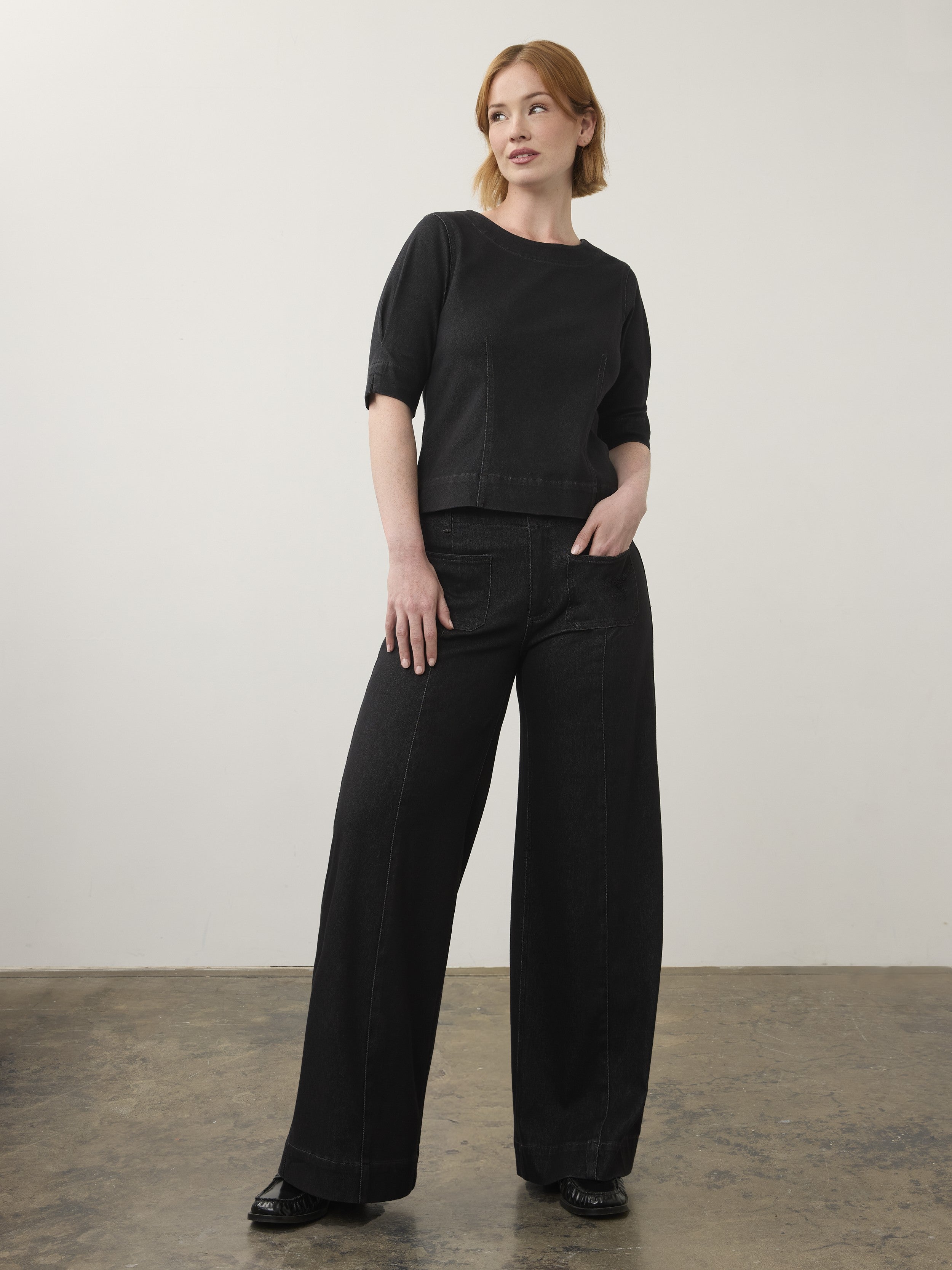 Individual standing indoors wearing a black short-sleeve top and wide-leg black denim pants with black shoes