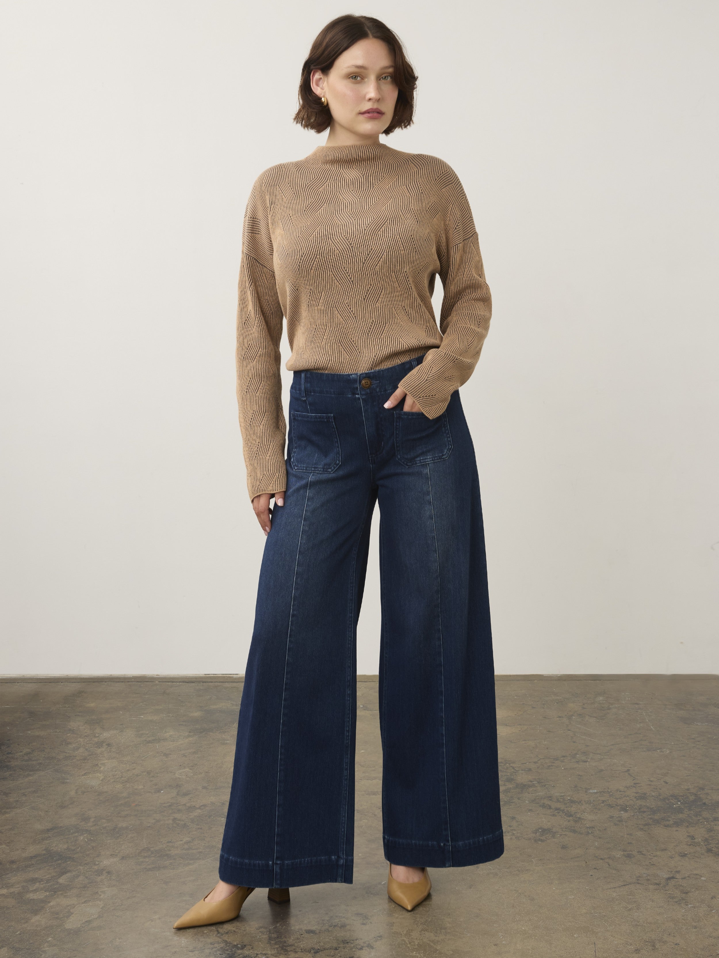 Individual in a beige textured sweater and wide-leg dark blue denim pants standing on a concrete floor against a plain wall.