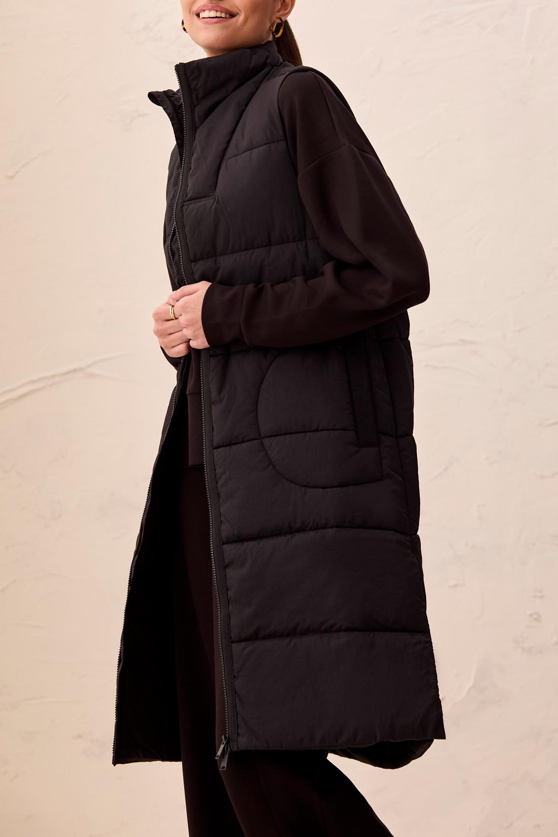 Long black quilted sleeveless vest zipped up over a black long-sleeve top against a beige background.