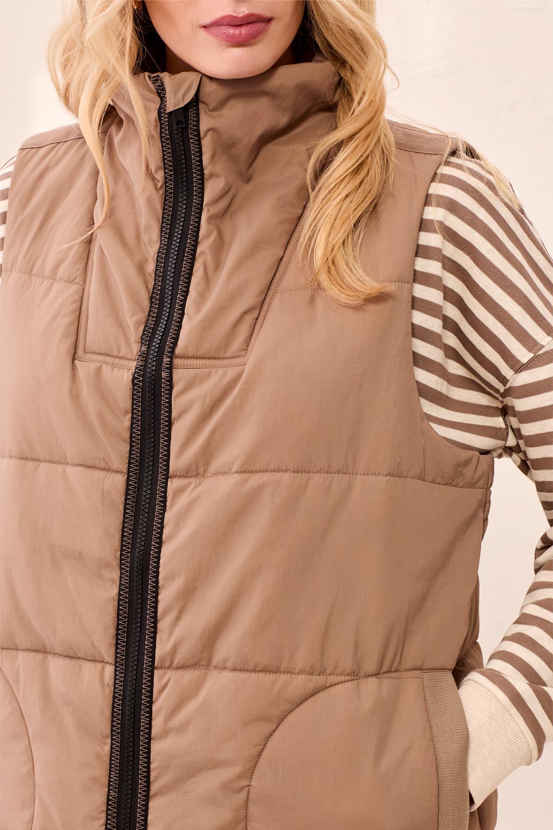 Close-up of a beige quilted vest with black zipper worn over a striped long-sleeve top with beige and white stripes.