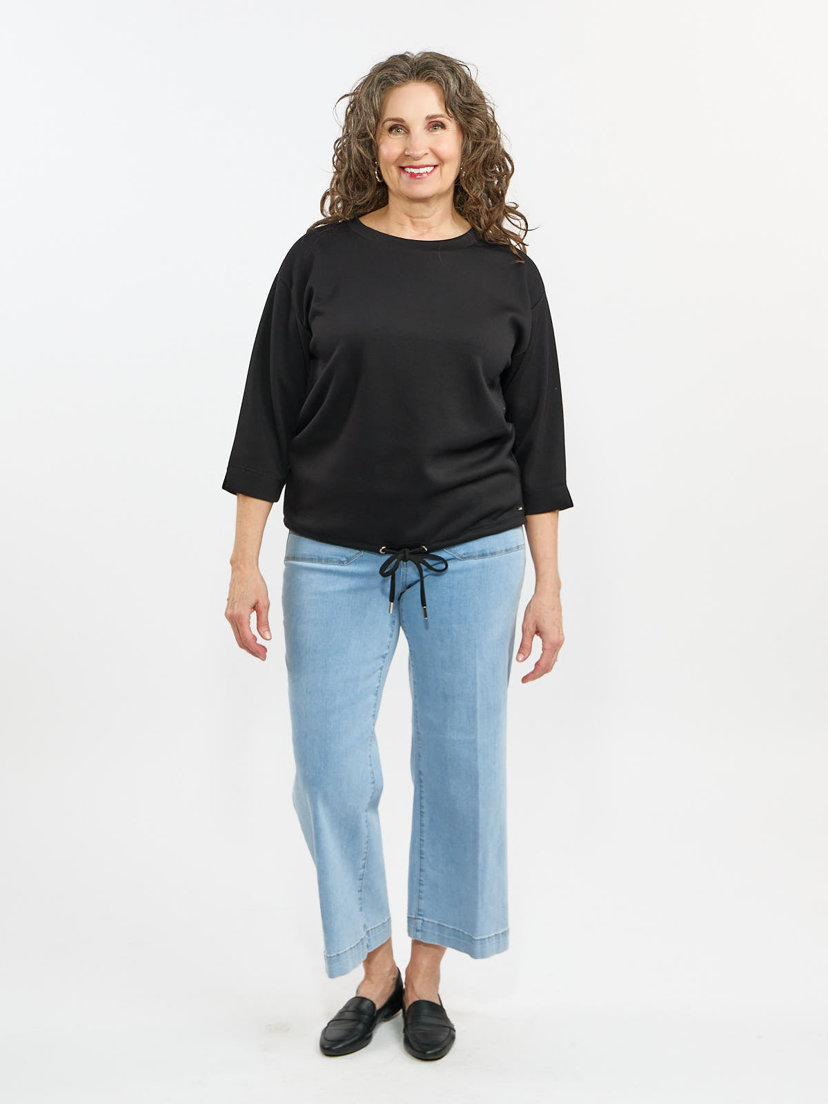 Model in a black long-sleeve top with drawstring hem, light blue wide-leg jeans, and black slip-on shoes.