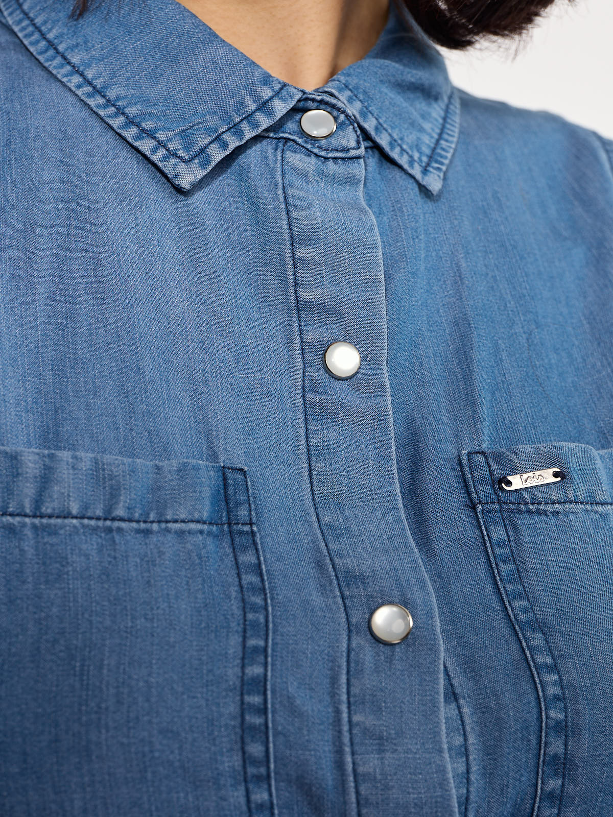 Close-up of a blue denim shirt with pearl snap buttons and two front chest pockets with stitching details