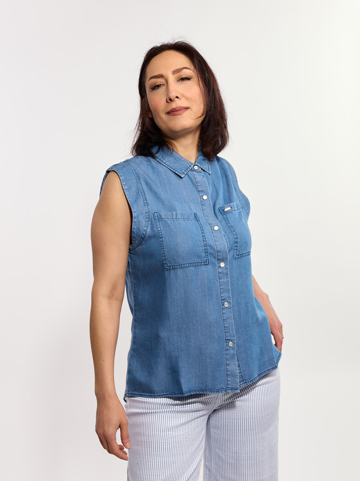 Sleeveless blue denim button-up shirt with front pockets paired with light striped pants against a white background.