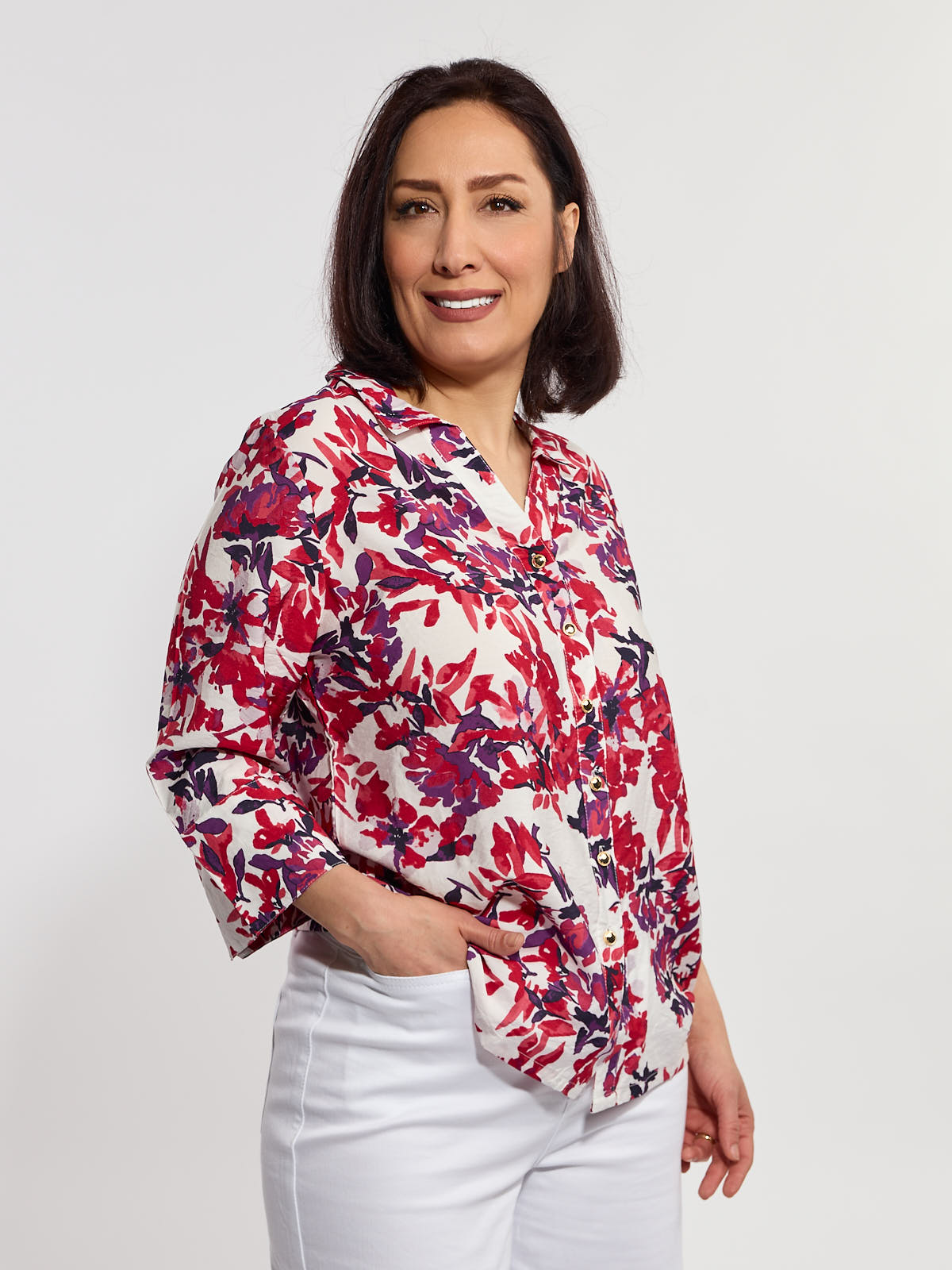 Woman in a red and purple floral button-up blouse with three-quarter sleeves and white pants against a light background