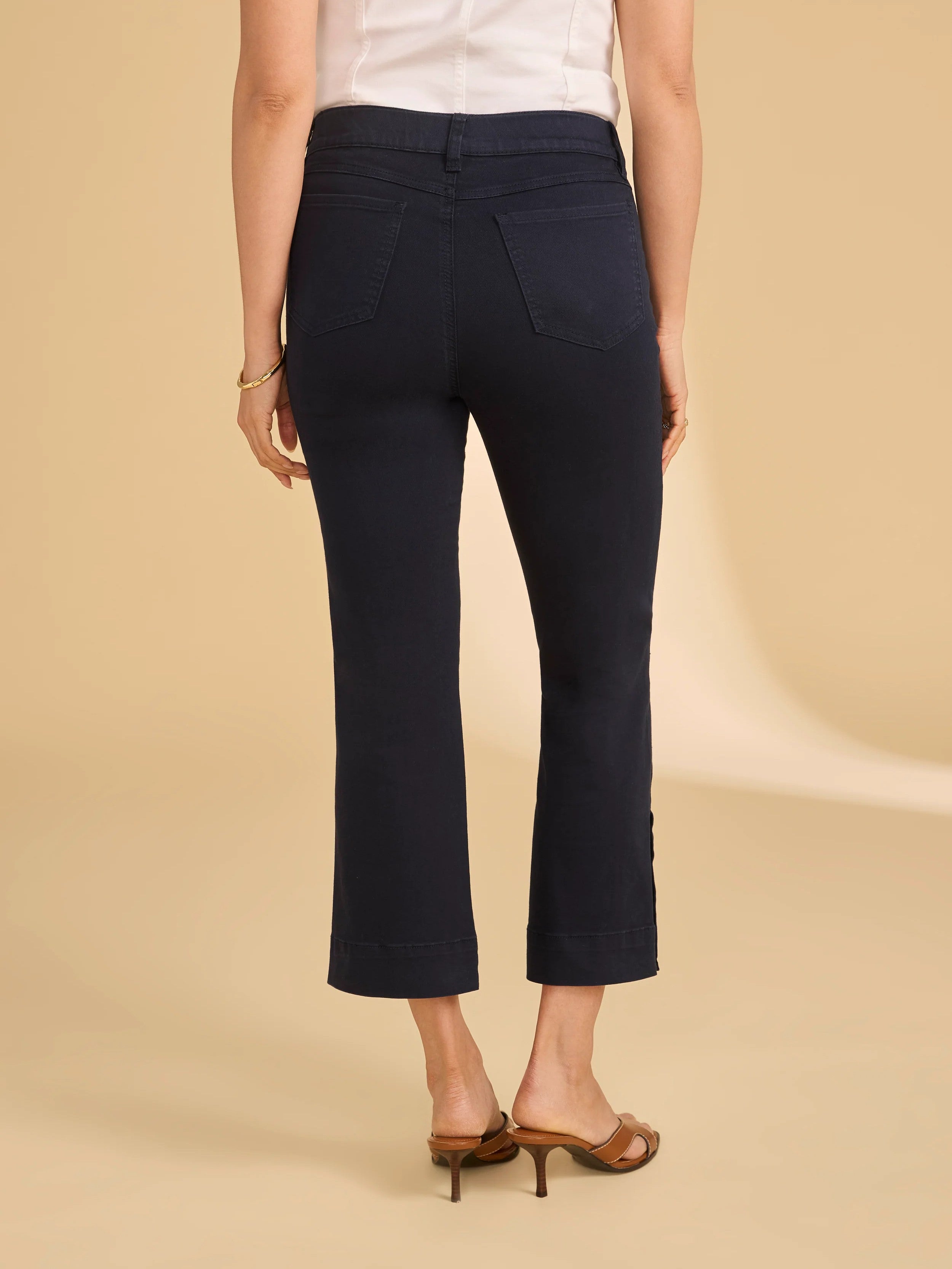 Back view of navy cropped pants with back pockets paired with brown heeled sandals on neutral background.