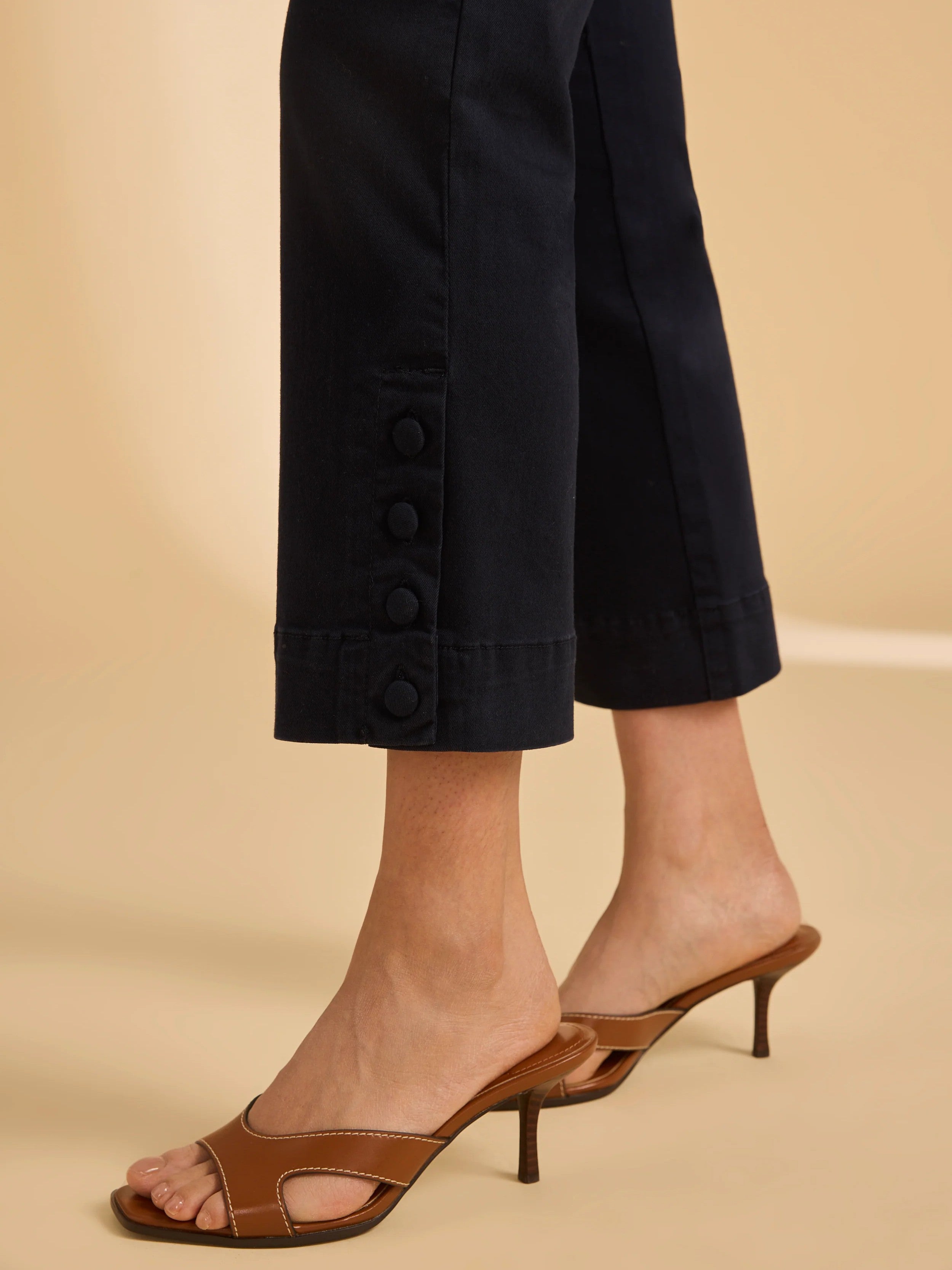 Close-up of cropped black pants featuring four fabric-covered buttons near the hem paired with brown heeled sandals.