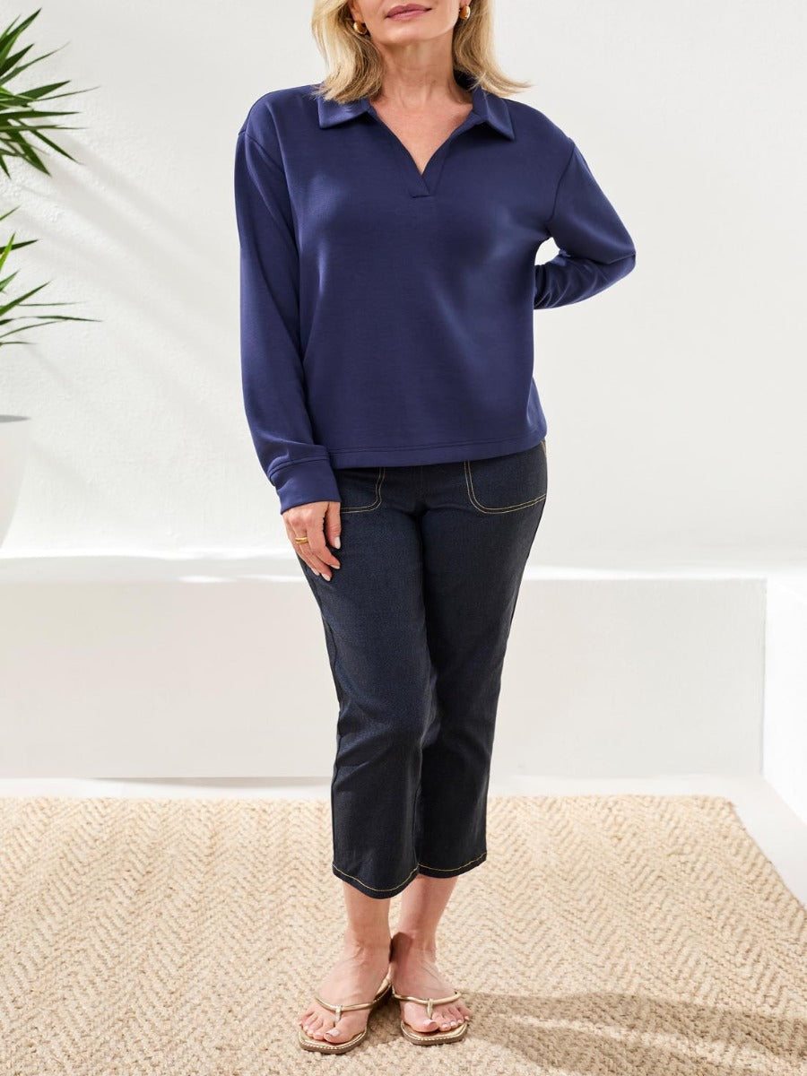 Light-skinned figure wearing a navy long-sleeve top with collar and dark cropped denim pants standing indoors on a woven rug.