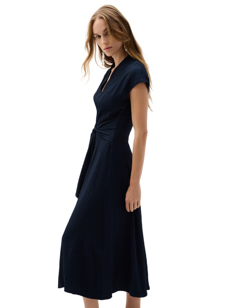 Model in a dark navy textured midi dress with short sleeves and a front waist tie on a white background