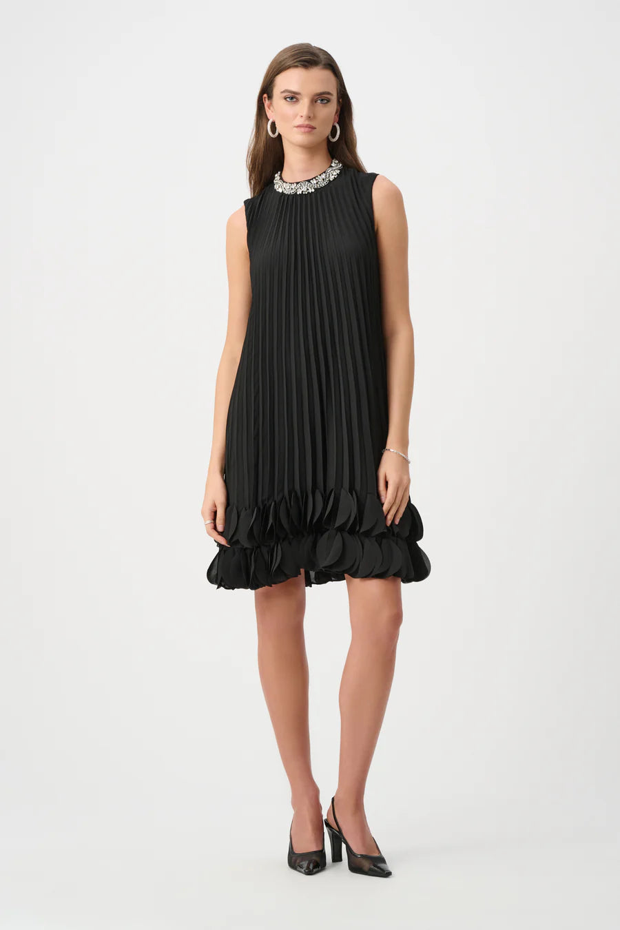 Sleeveless black pleated dress with embellished neckline and decorative ruffle hem paired with black heels and silver hoop earrings.
