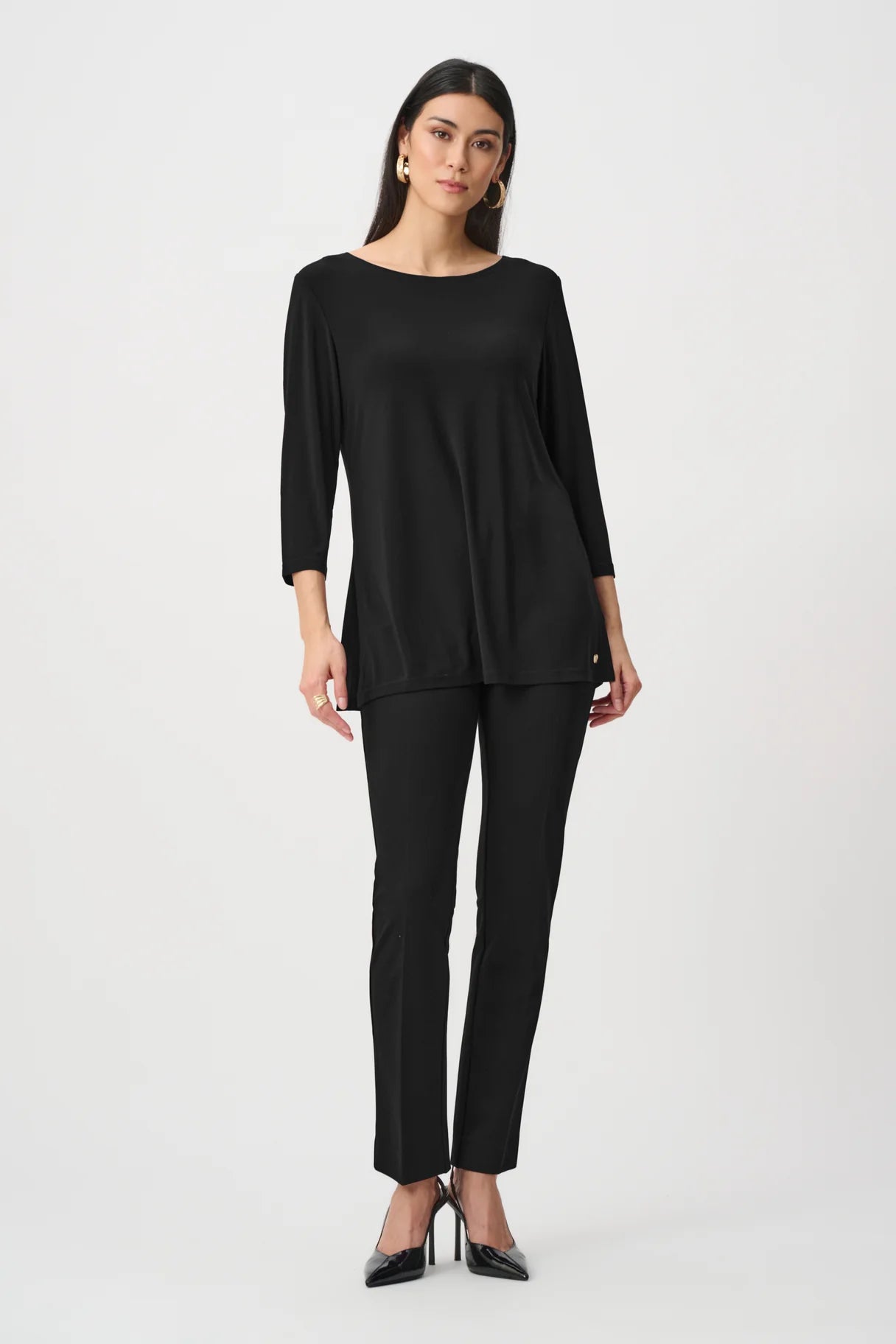 Model standing in a black long-sleeve top and black fitted pants with black pointed heels against a white background