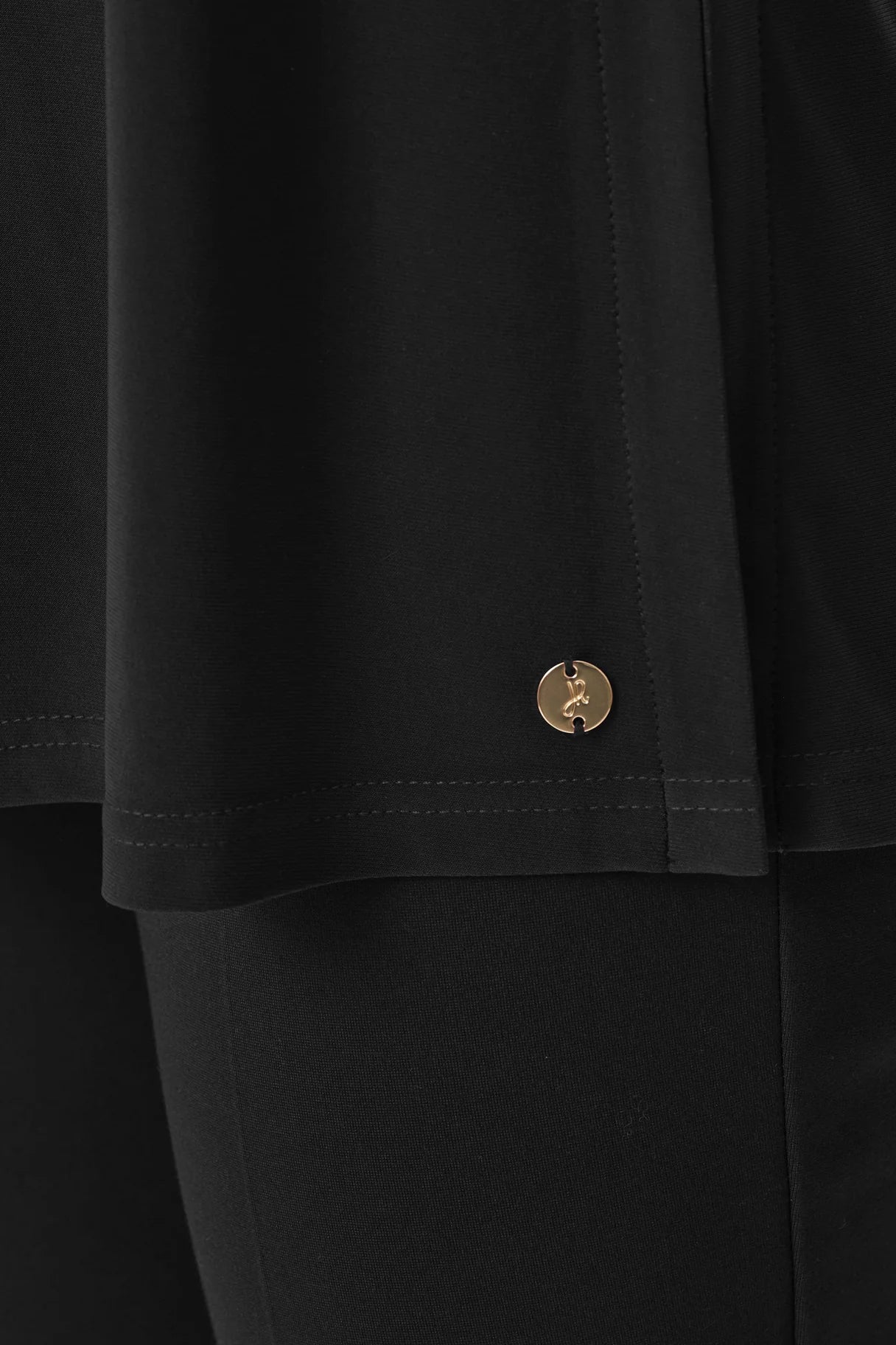 Close-up of black outerwear hem with a small round gold metallic logo detail stitched near the edge.