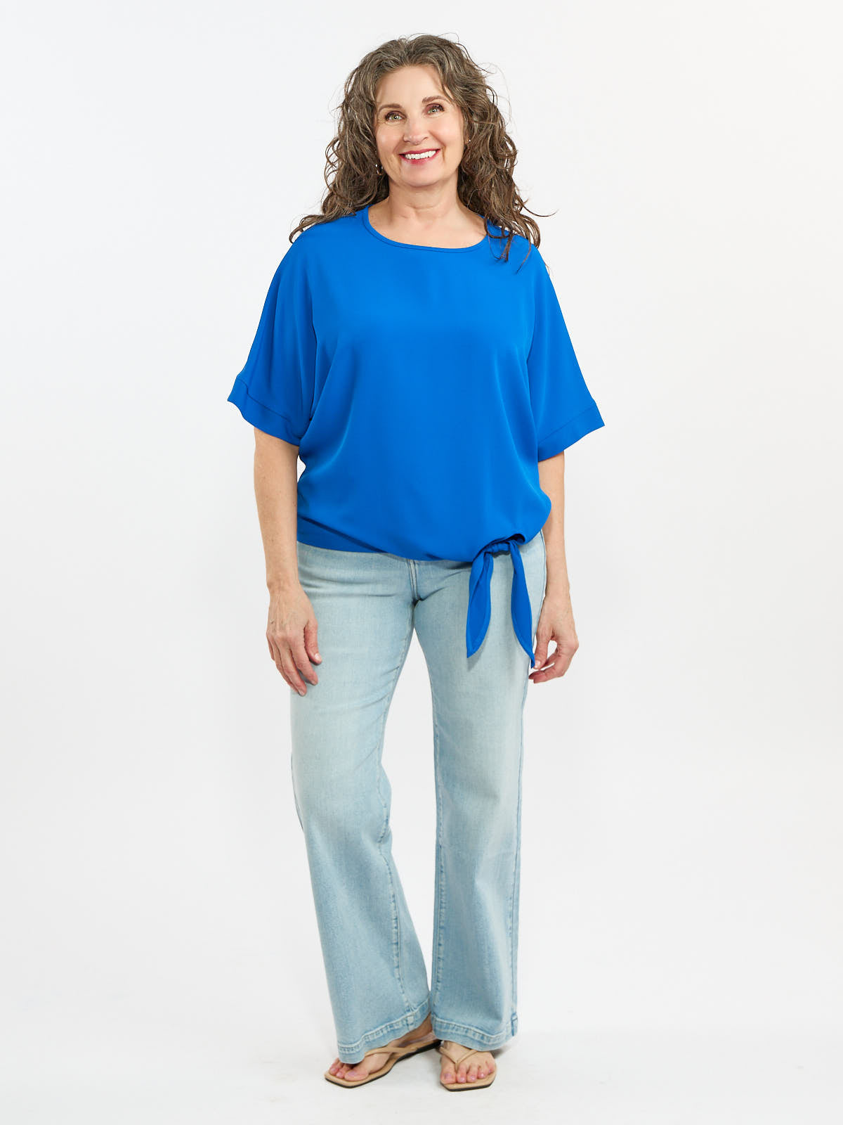 Blue short-sleeve blouse with side tie paired with light wash denim jeans and brown sandals on white background.