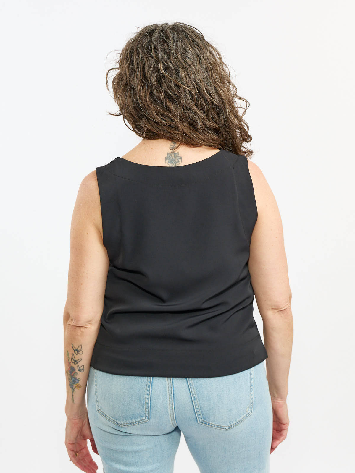 Back view of sleeveless black top paired with light blue denim jeans on a person with curly hair and visible tattoos.