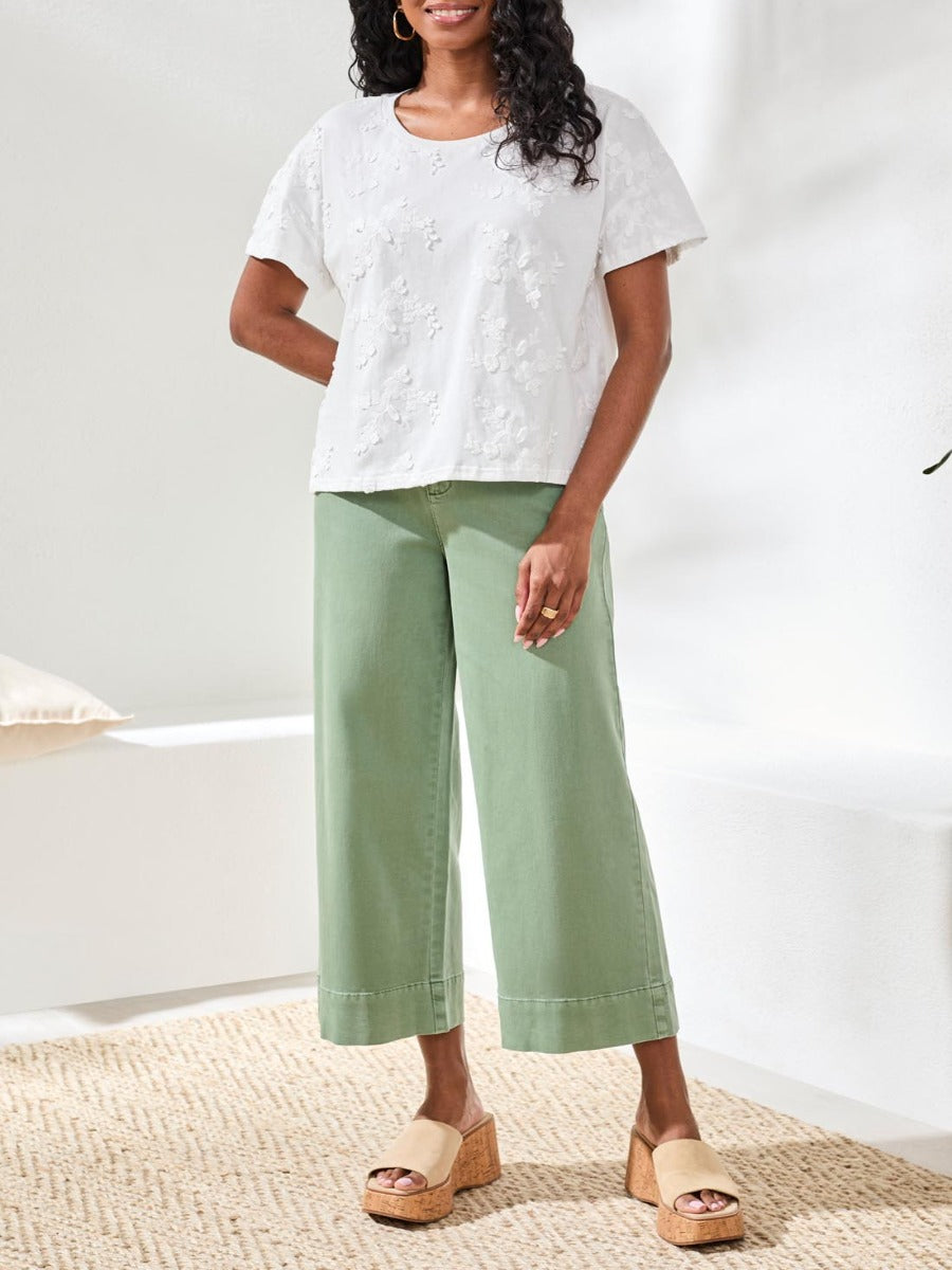 Person stands indoors wearing a white floral textured top, wide-leg sage green pants, and beige platform sandals.