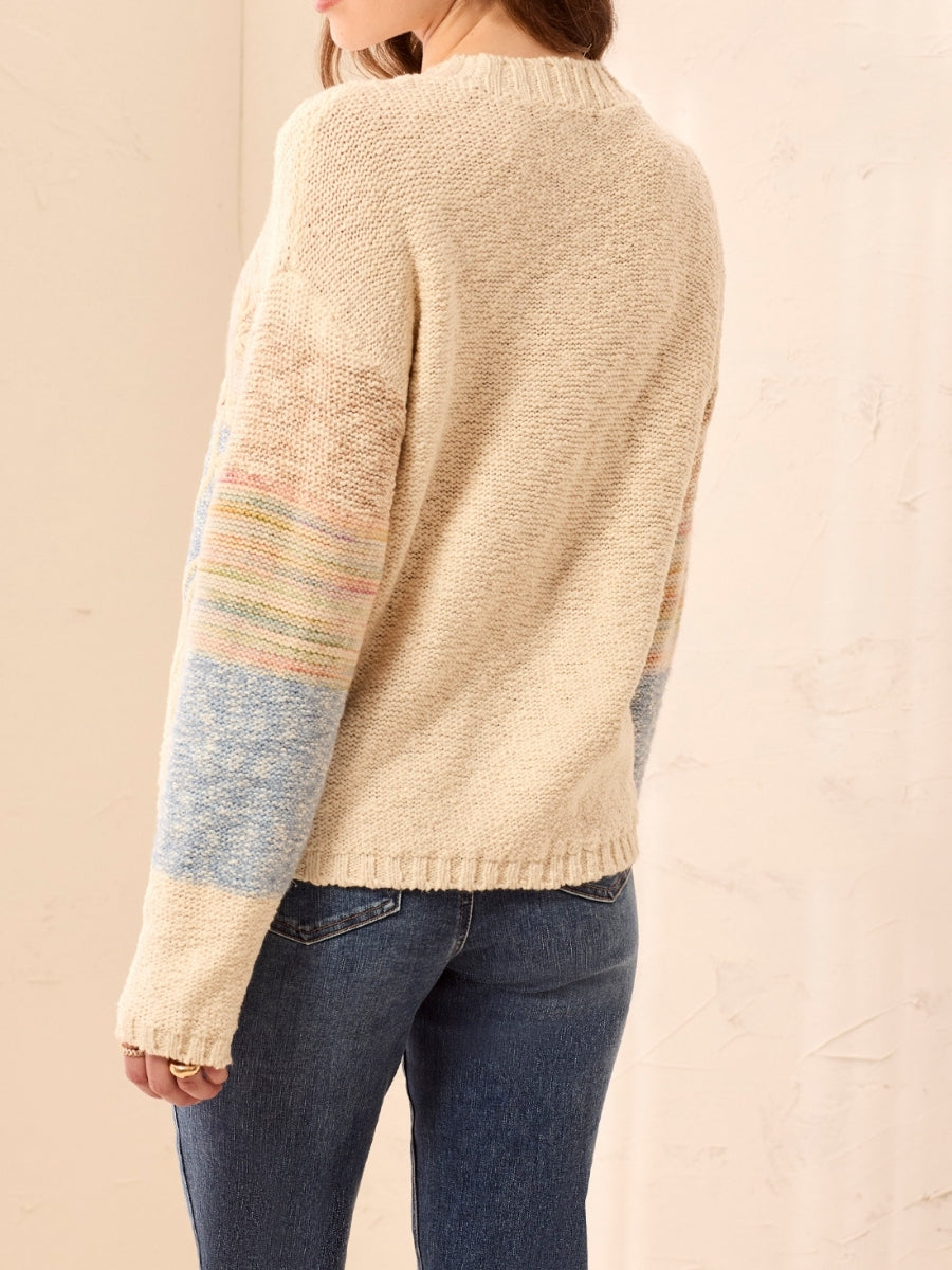 Cream knit sweater with multicolored striped sleeves paired with dark denim jeans against a neutral background.