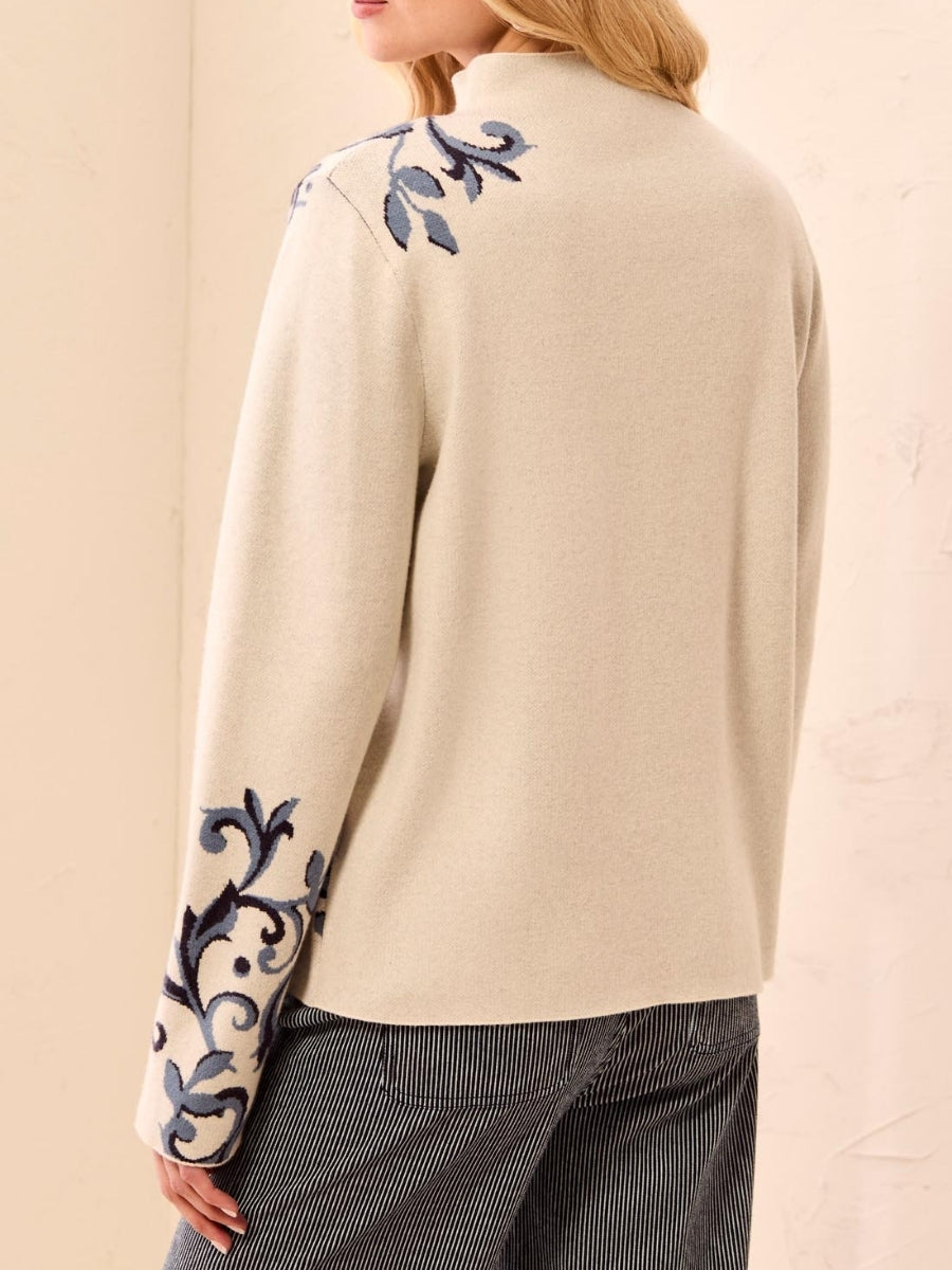 Cream-colored sweater with blue floral embroidery on shoulder and sleeve, paired with dark pinstriped pants.