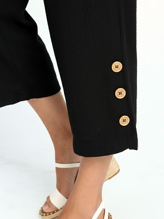 Close-up of black cropped pants with three wooden buttons near the hem and white strappy wedge sandals.