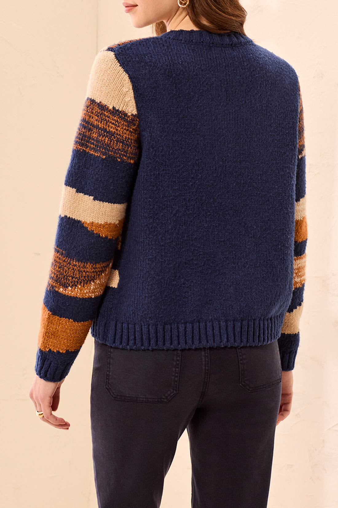 Back view of a navy blue sweater with beige, orange, and brown striped sleeves paired with dark pants.