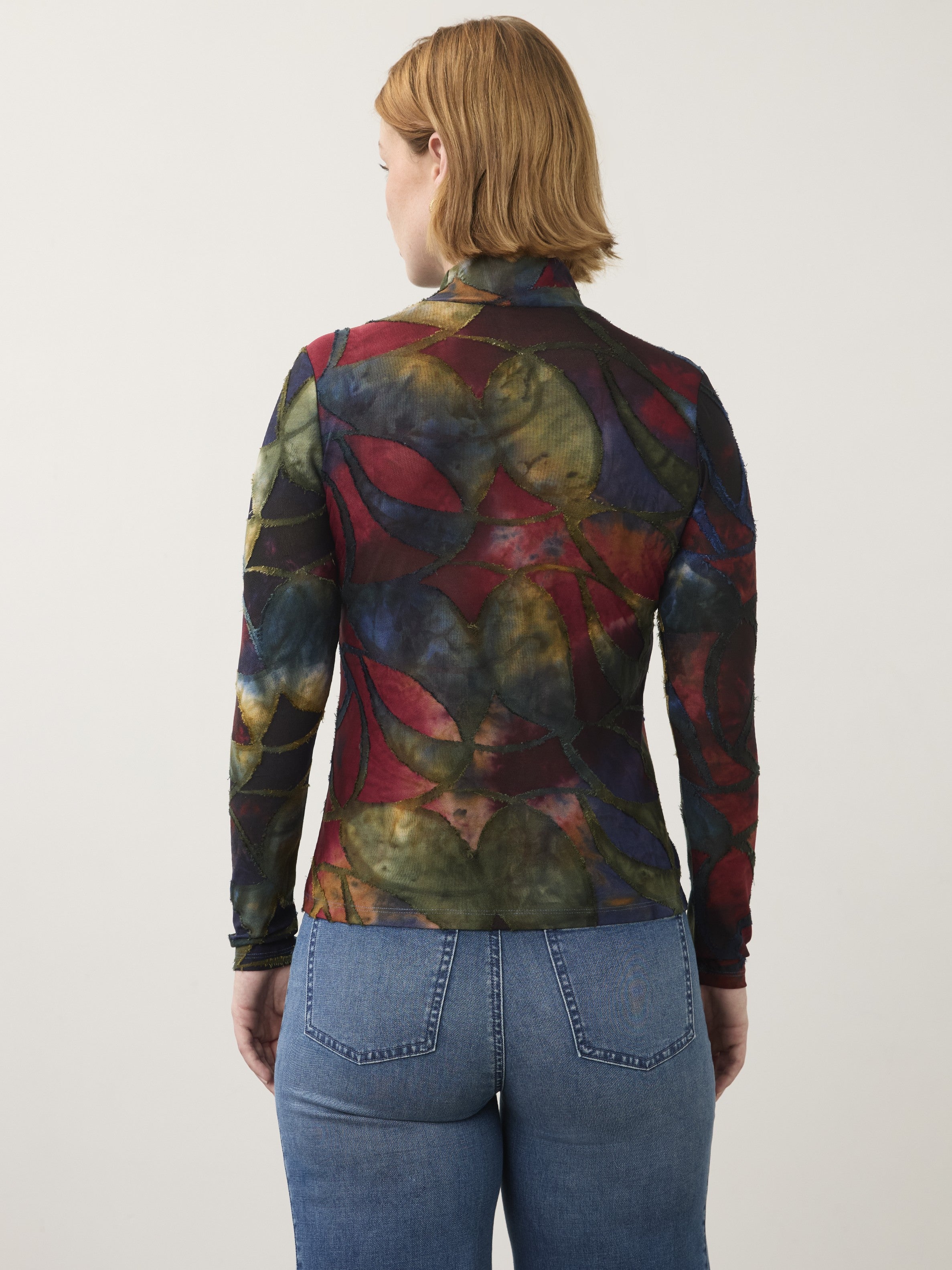 Back view of a long-sleeve, multicolor patterned top paired with blue denim jeans on a neutral background.