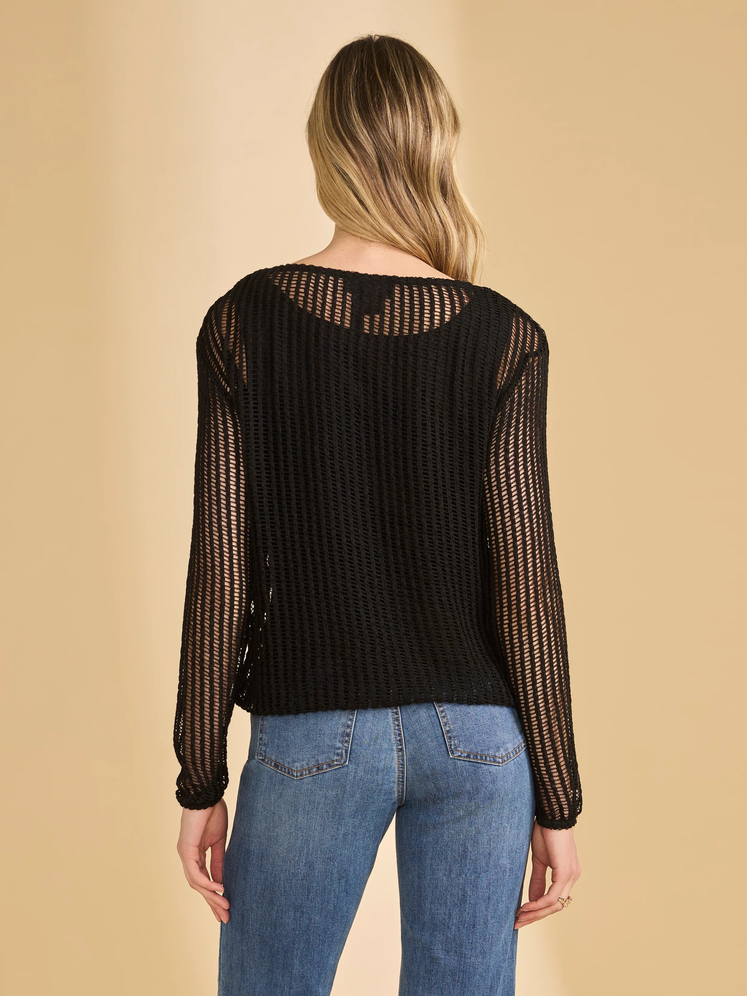Black long-sleeve sheer knit top with vertical openwork pattern paired with blue denim jeans against a beige background.