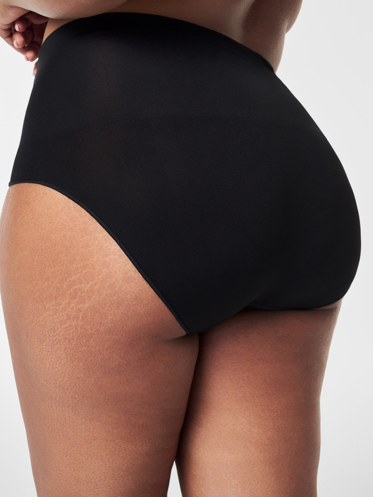 Rear view of black SPANX EcoCare Seamless Brief by Spanx on skin of a tan model with stretch texture.