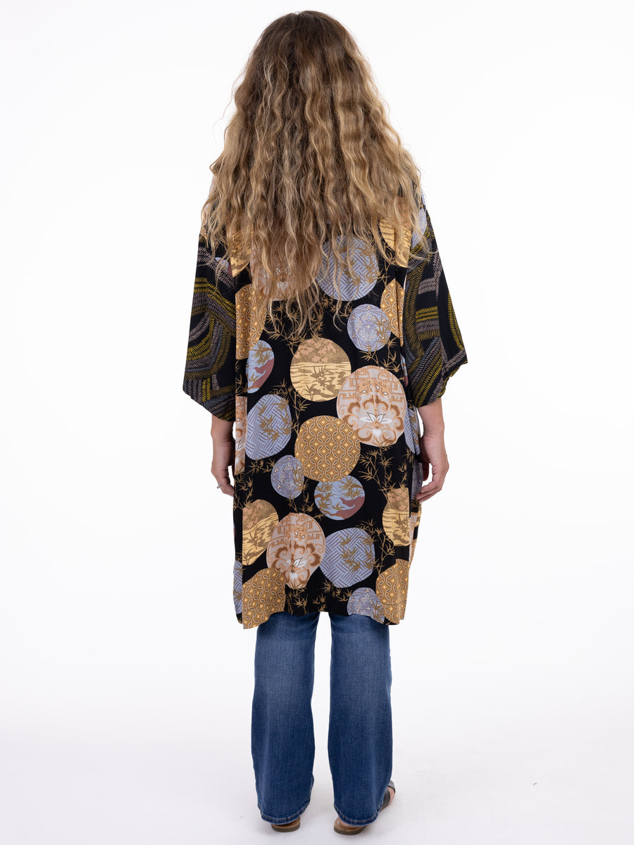 Person with long curly hair wearing a patterned kimono-style top and blue jeans, facing away from the camera.