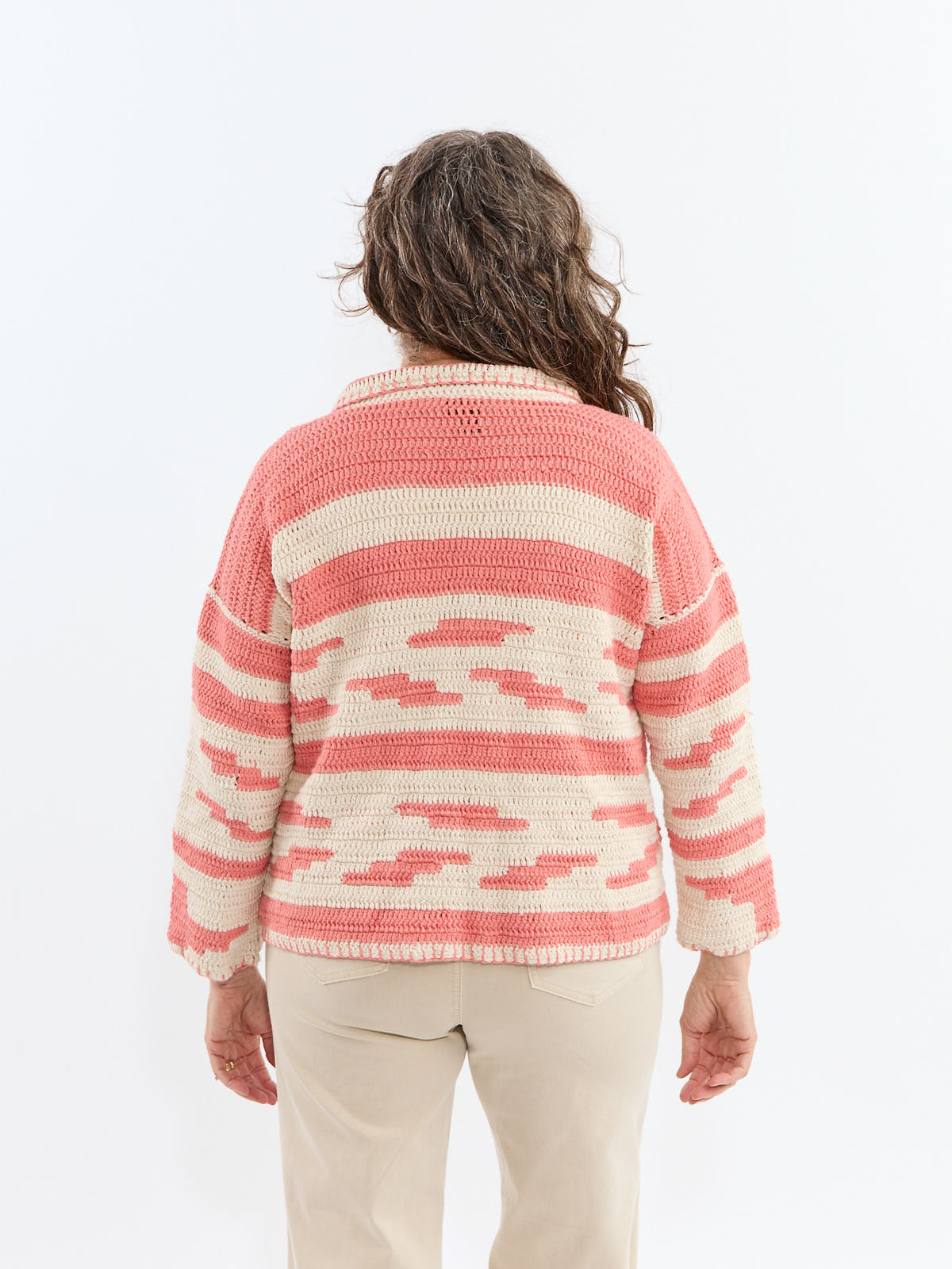 Back view of a person wearing a coral and cream striped knitted sweater with beige pants against a white background.