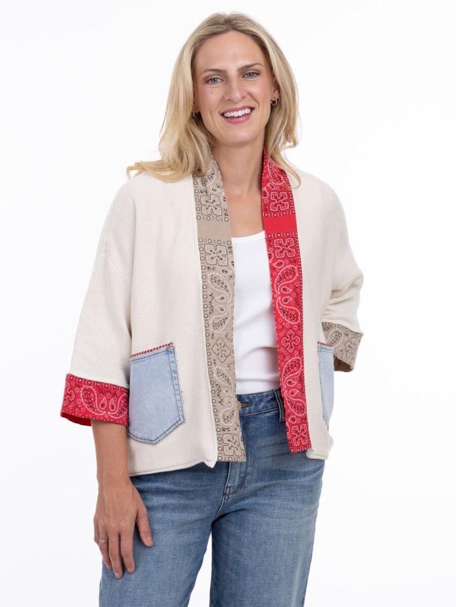 Smiling person wearing cream knit open cardigan with red and beige patterned trim and denim patch pockets over white top and jeans