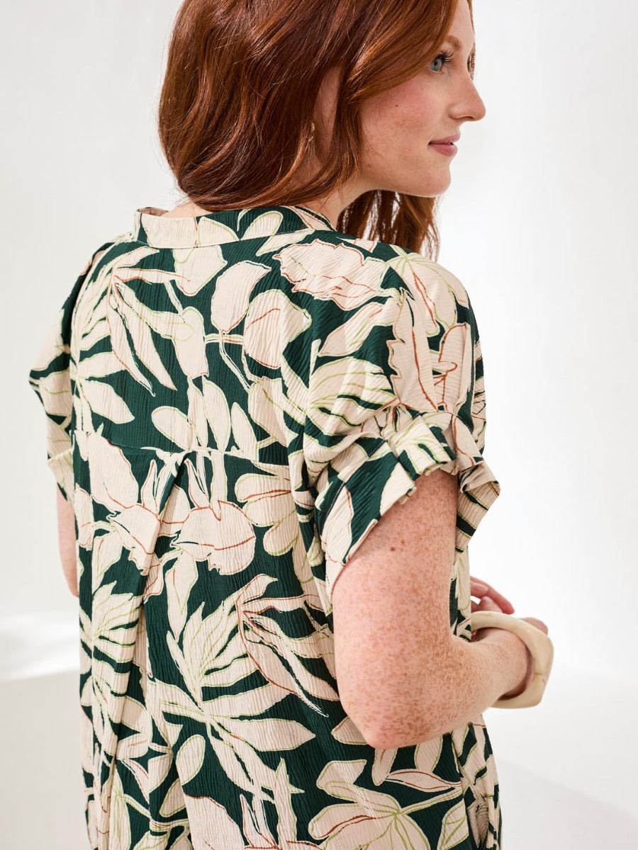 Side view of a person with red hair wearing a green and beige leaf-patterned short-sleeve top and a cream bracelet.