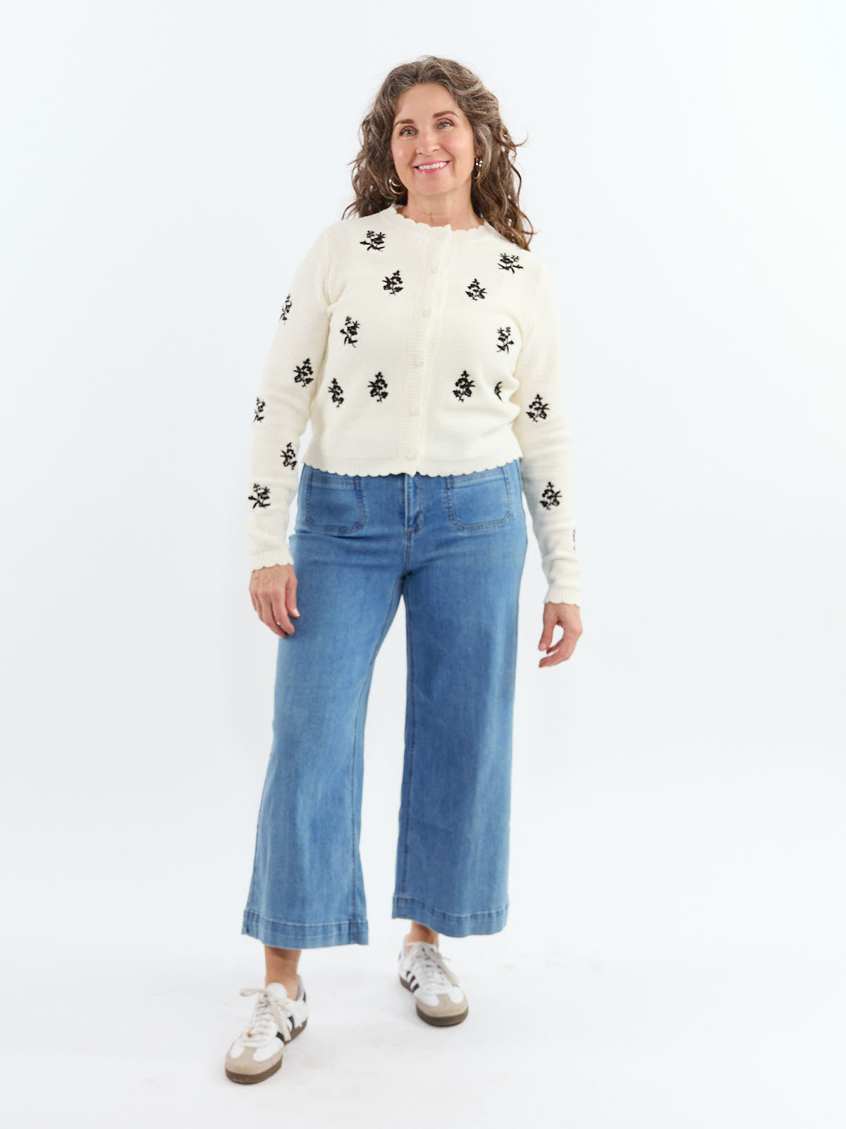 Person wearing a cream cardigan with black floral designs, blue wide-leg jeans, and white sneakers against a white background
