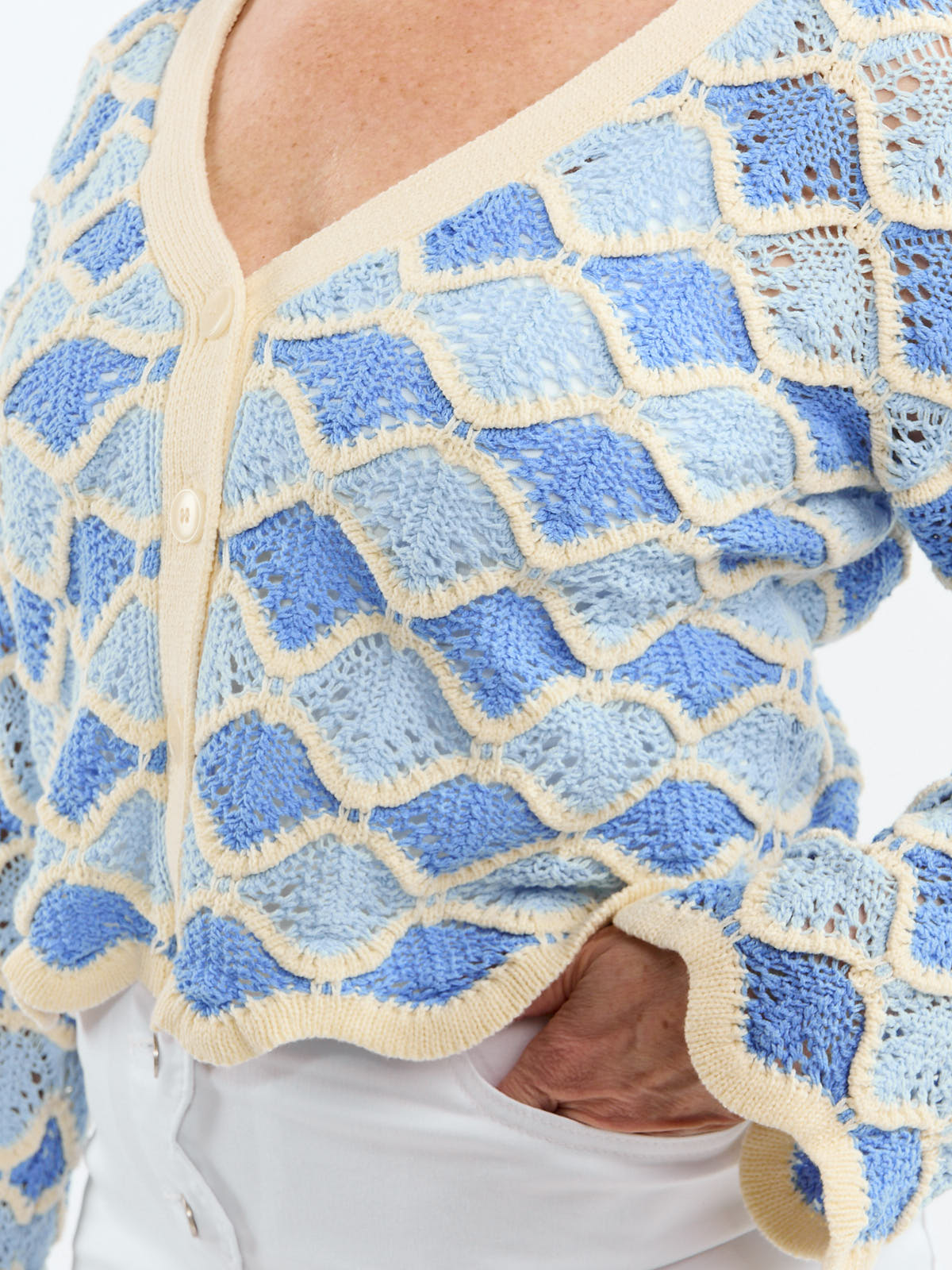 Close-up of a textured blue and cream knit sweater with a scalloped edge and buttons over white pants.