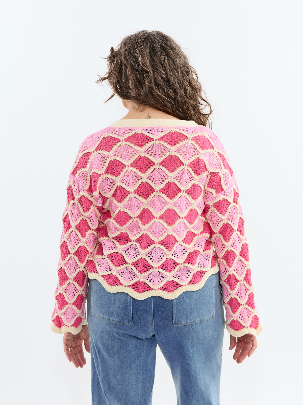 Open-knit pink and red patterned sweater with scalloped edges worn with blue jeans viewed from the back